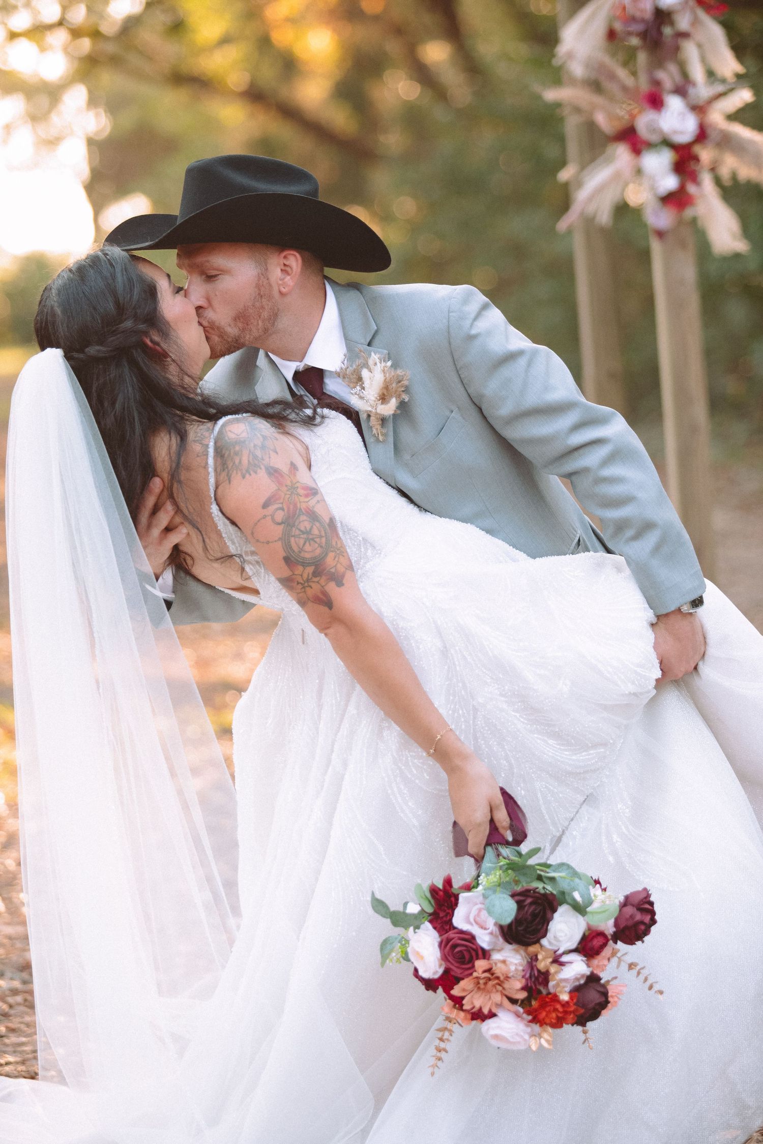Groom in cowboy hat dips bride in white lace gown and veil for a kiss, holding a burgundy bouquet outdoors.