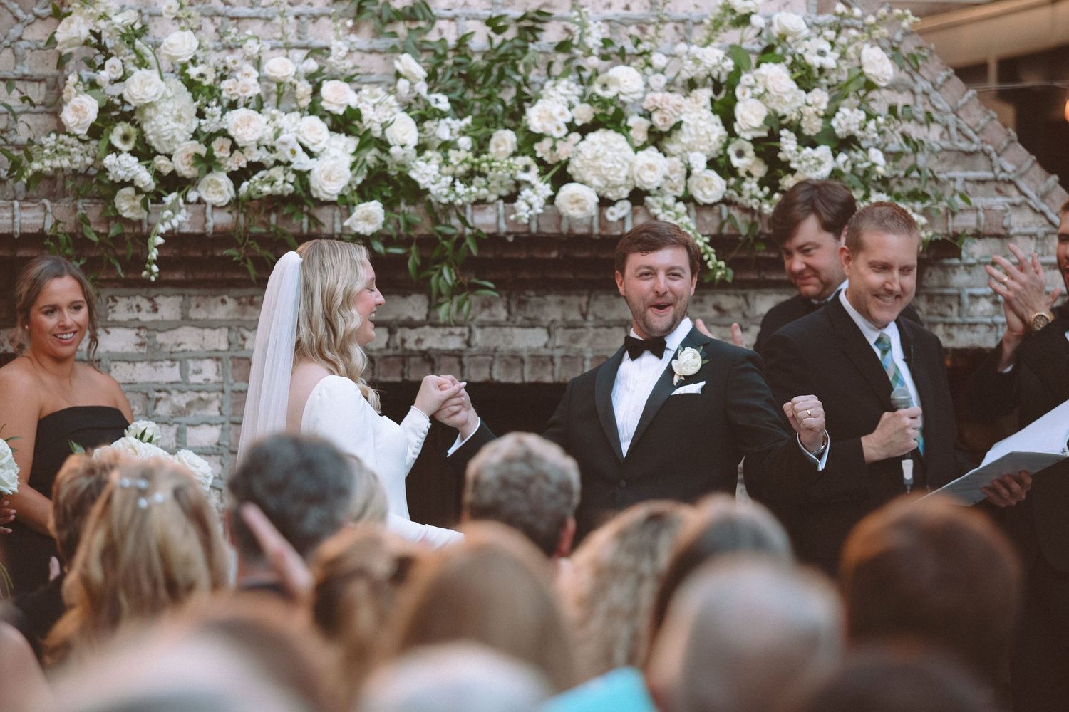 Joyful bride and groom celebrate at outdoor wedding ceremony with white floral arch over brick fireplace.