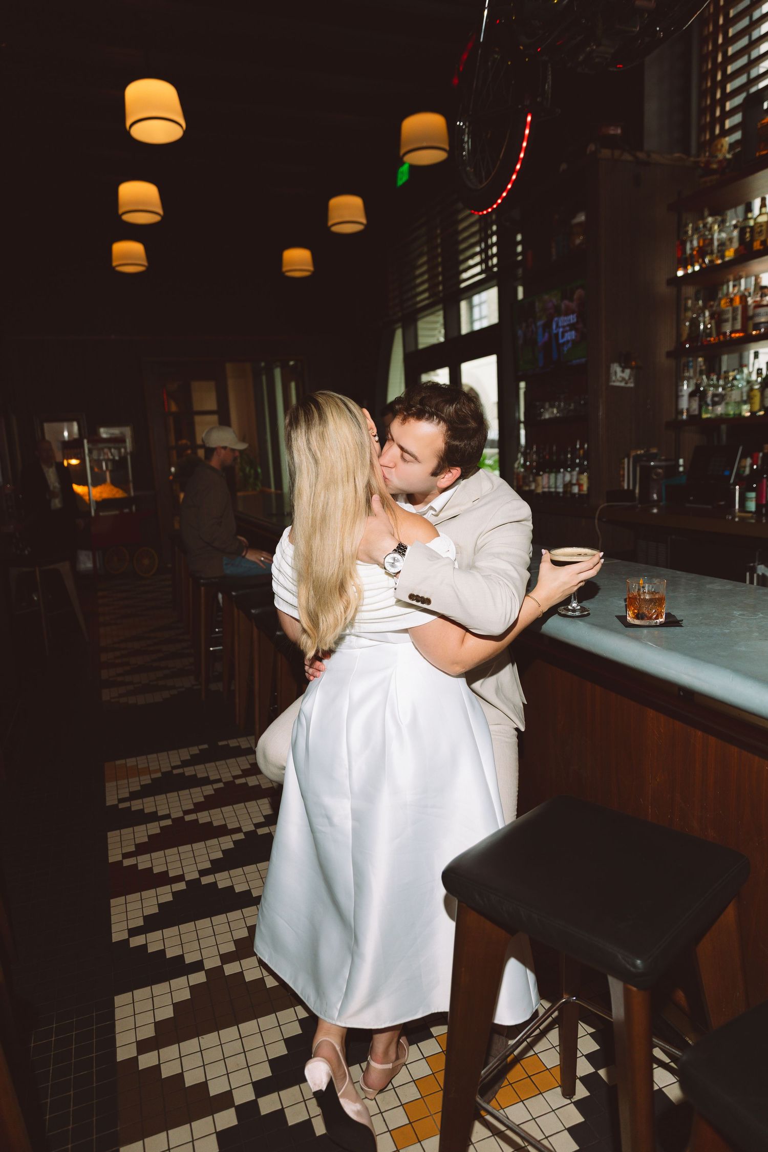 Couple sharing a romantic kiss at a dimly lit bar with warm pendant lights and a whiskey glass nearby.