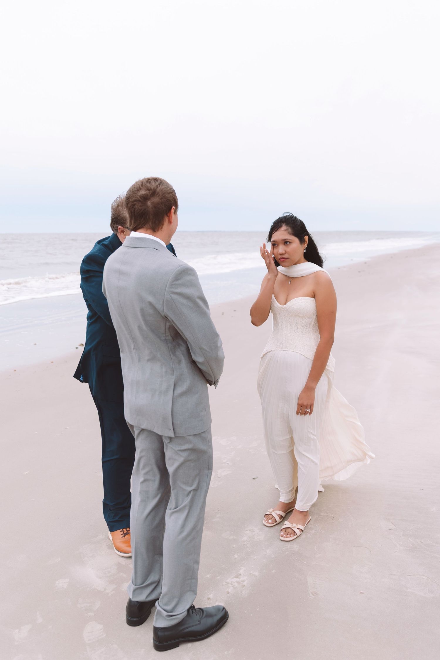 Emotional bride wiping tears during a beach wedding ceremony with groom and officiant on overcast day.