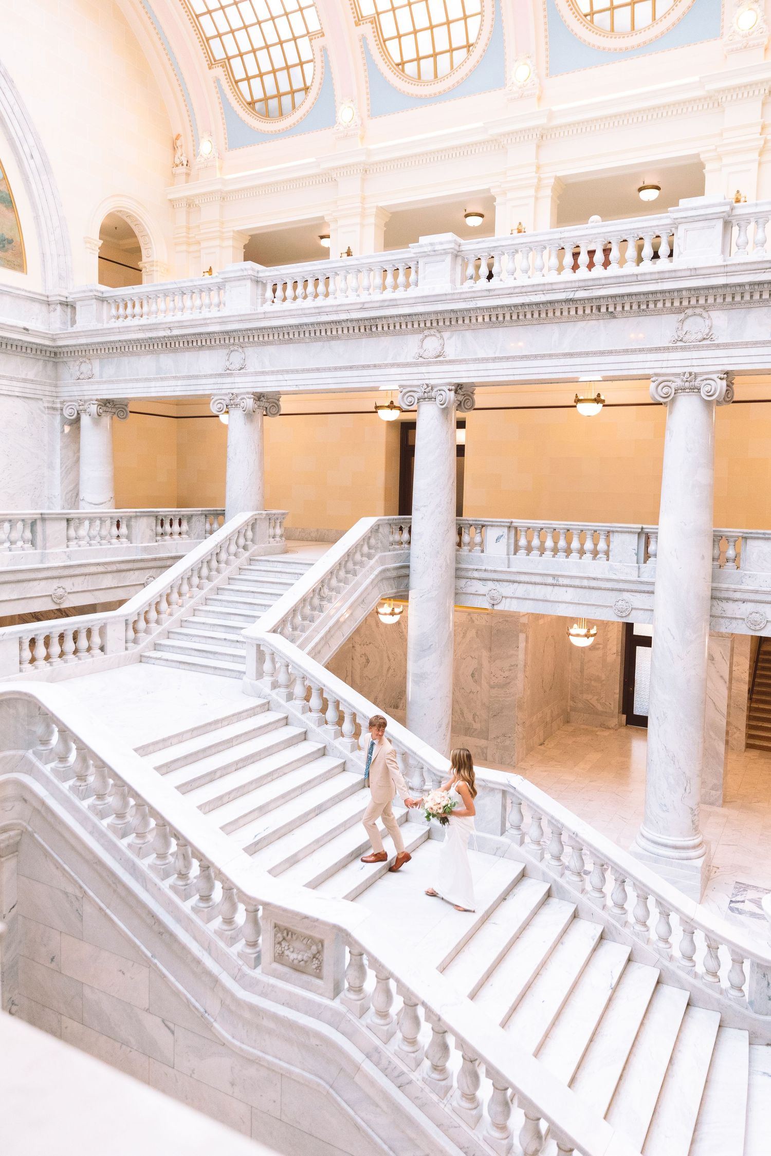 Wedding couple walks grand marble staircase inside elegant neoclassical capitol building.