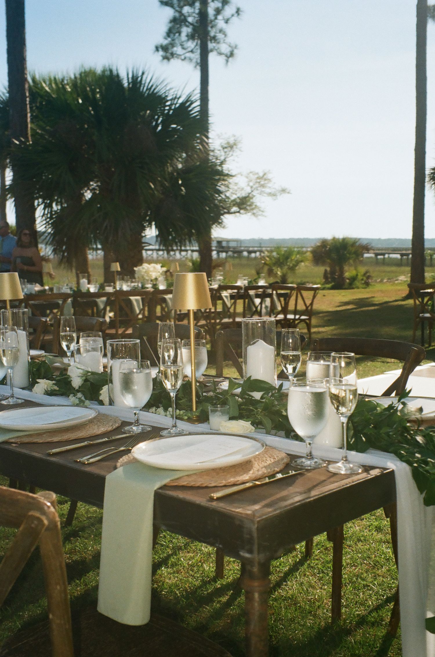 Outdoor waterfront wedding reception tablescape with rustic wood tables, gold lamps, and lush greenery runner.