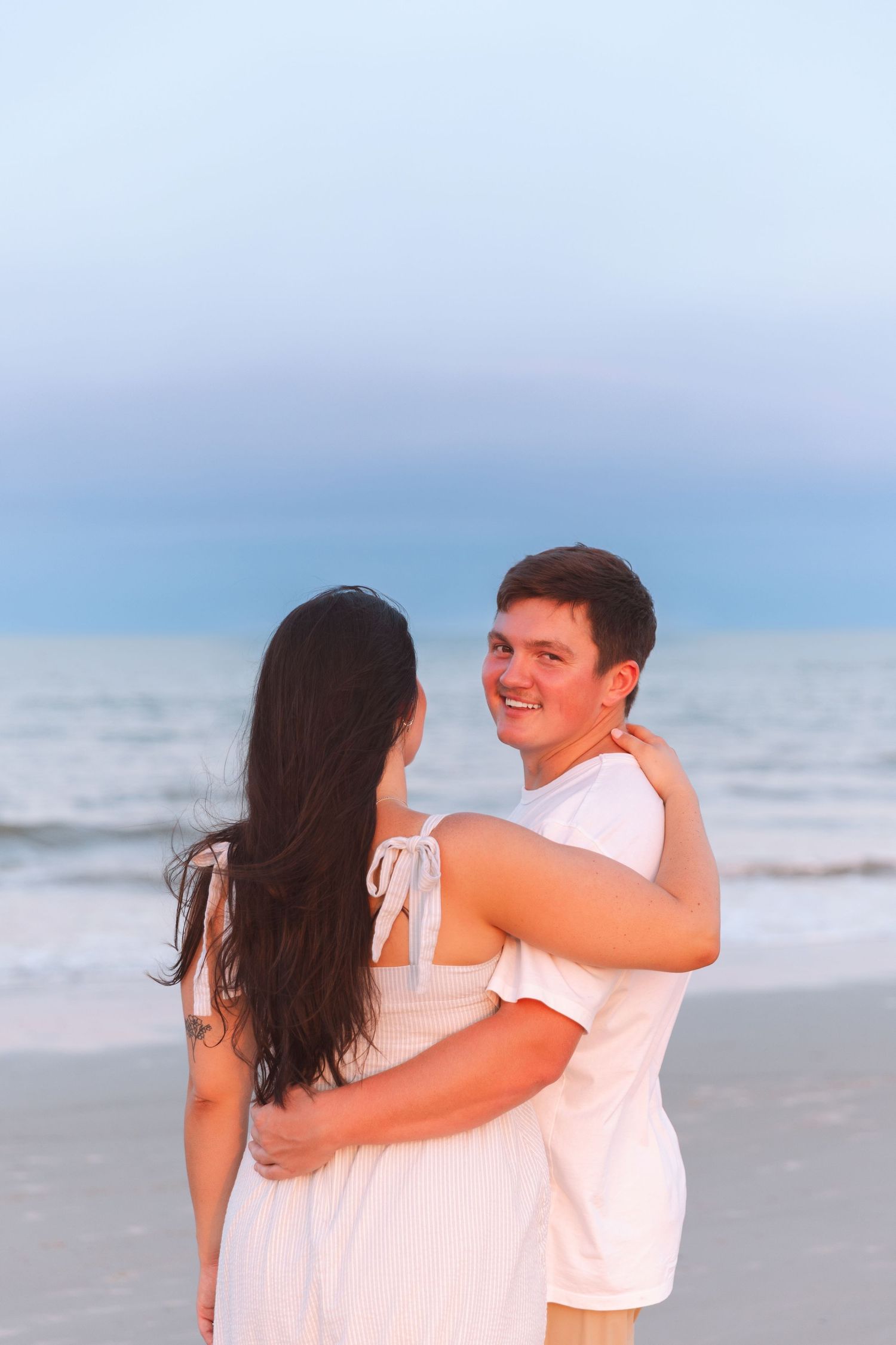 A smiling couple embraces on a beach at dusk, both wearing white outfits.