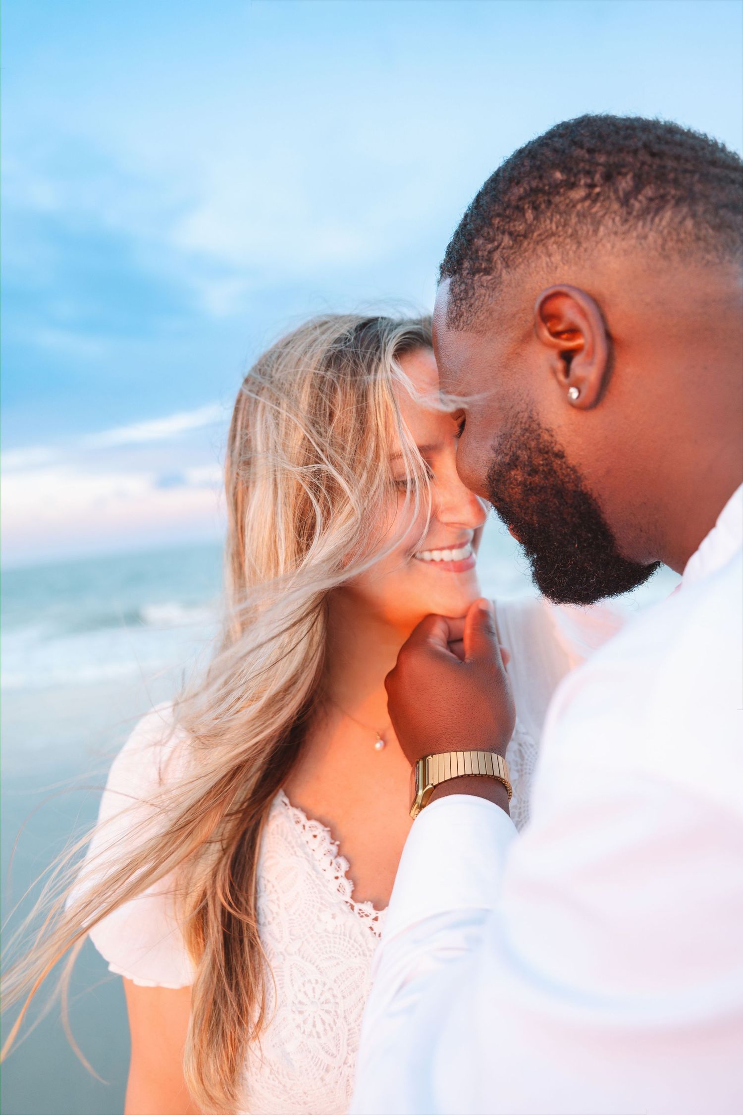Romantic interracial couple sharing a tender moment on a beach at sunset, man in white shirt gently holding womans chin.