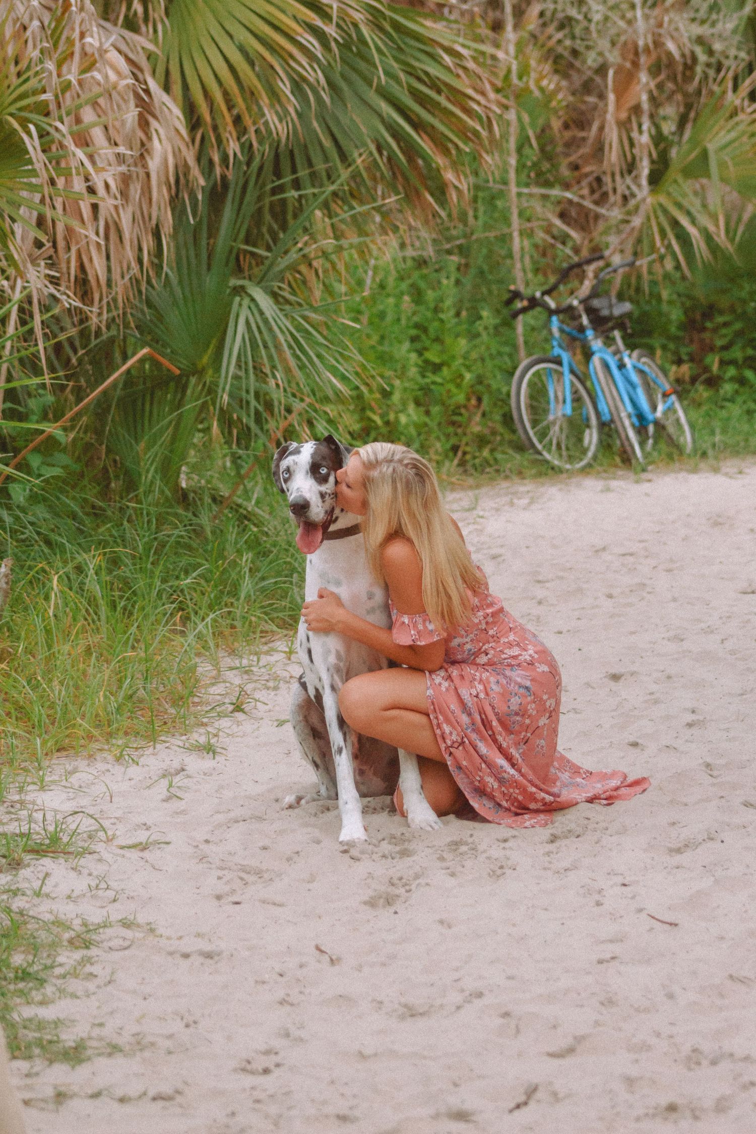 Blonde woman in floral dress kneeling and hugging a Great Dane dog on a sandy beach path.