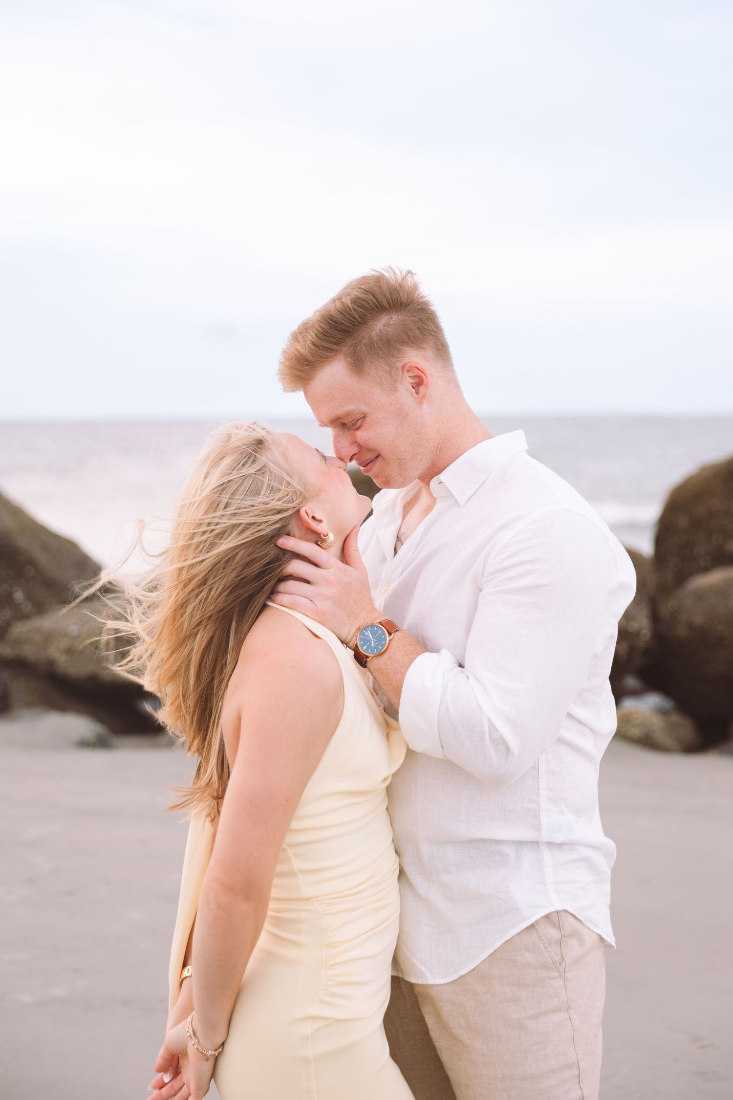 Romantic couple almost kissing on a beach with rocks, man in white shirt holding womans face.