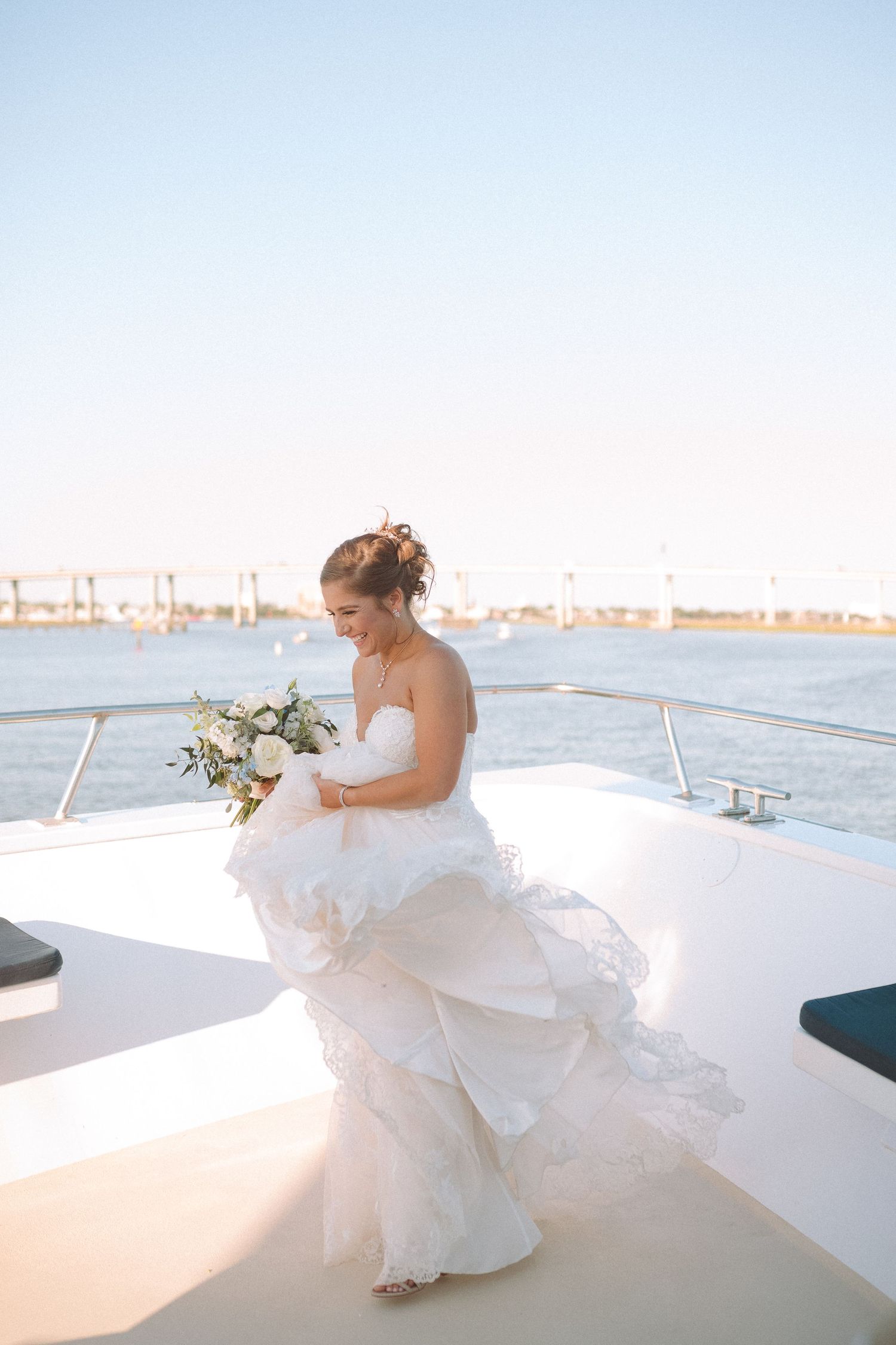 Radiant bride in strapless lace wedding gown holding bouquet on a yacht deck at sunset.