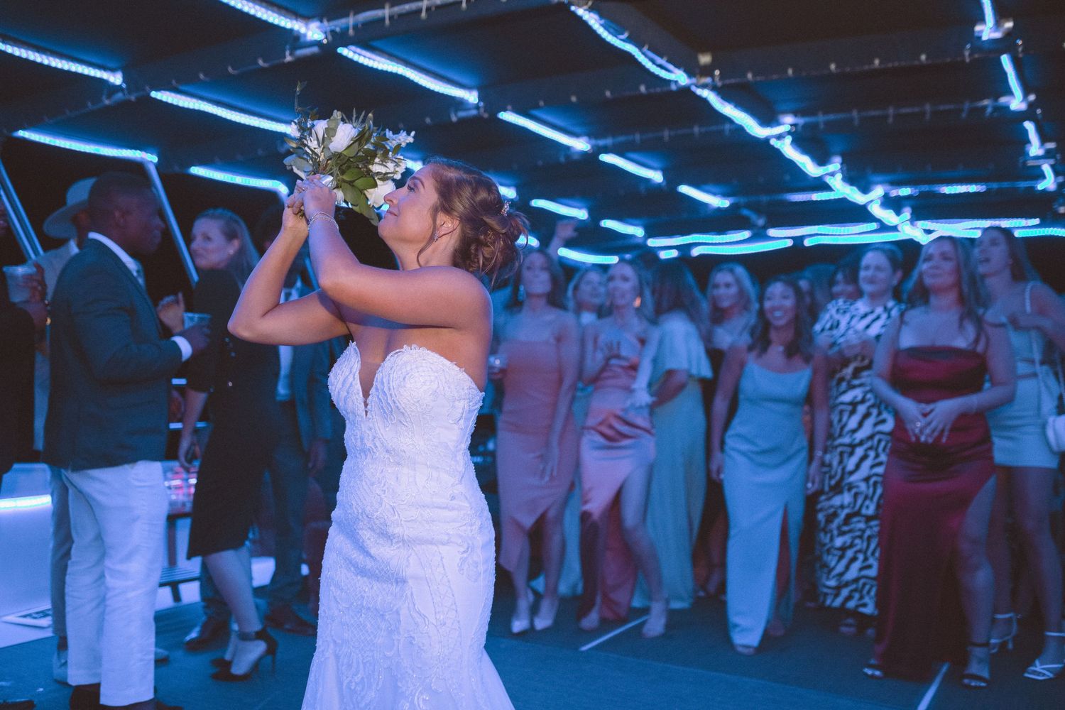 Bride in white lace gown tossing bouquet to excited wedding guests at reception.