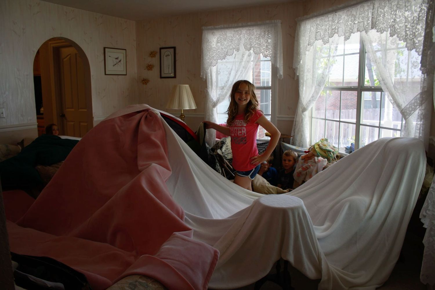 Young girl standing proudly beside a large homemade blanket fort built in a living room.