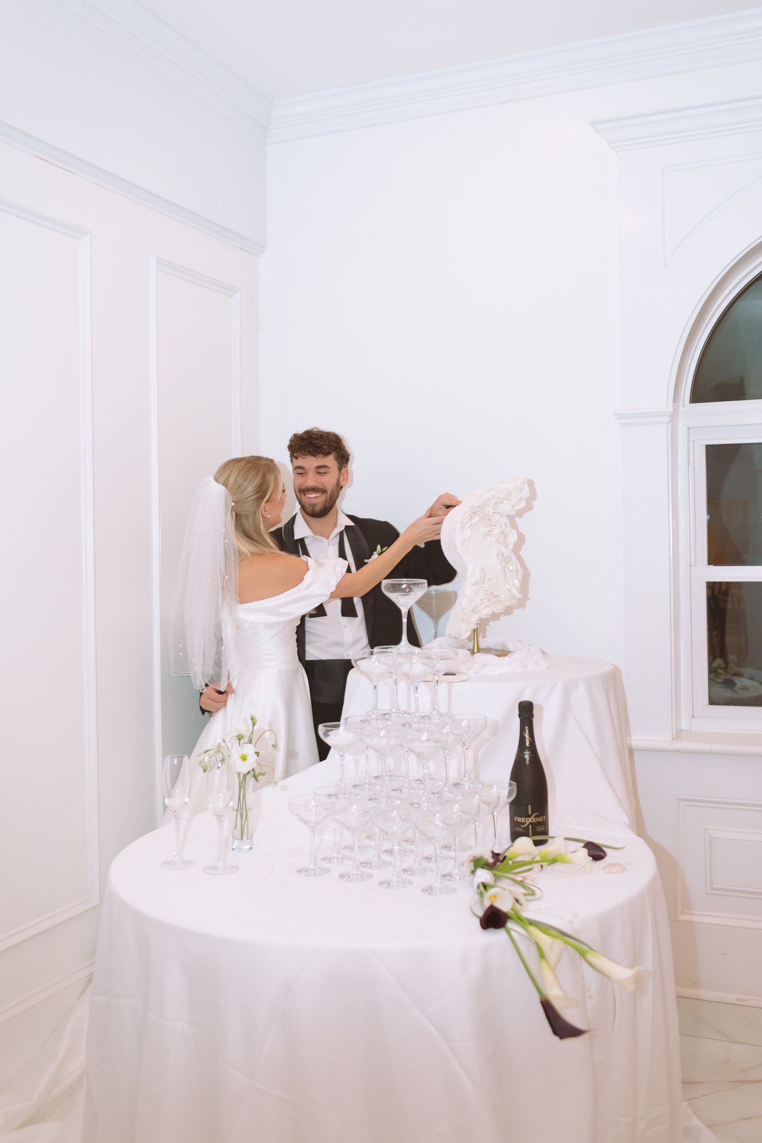 Smiling bride and groom interacting with a decorative sculpture beside a champagne tower at their reception.