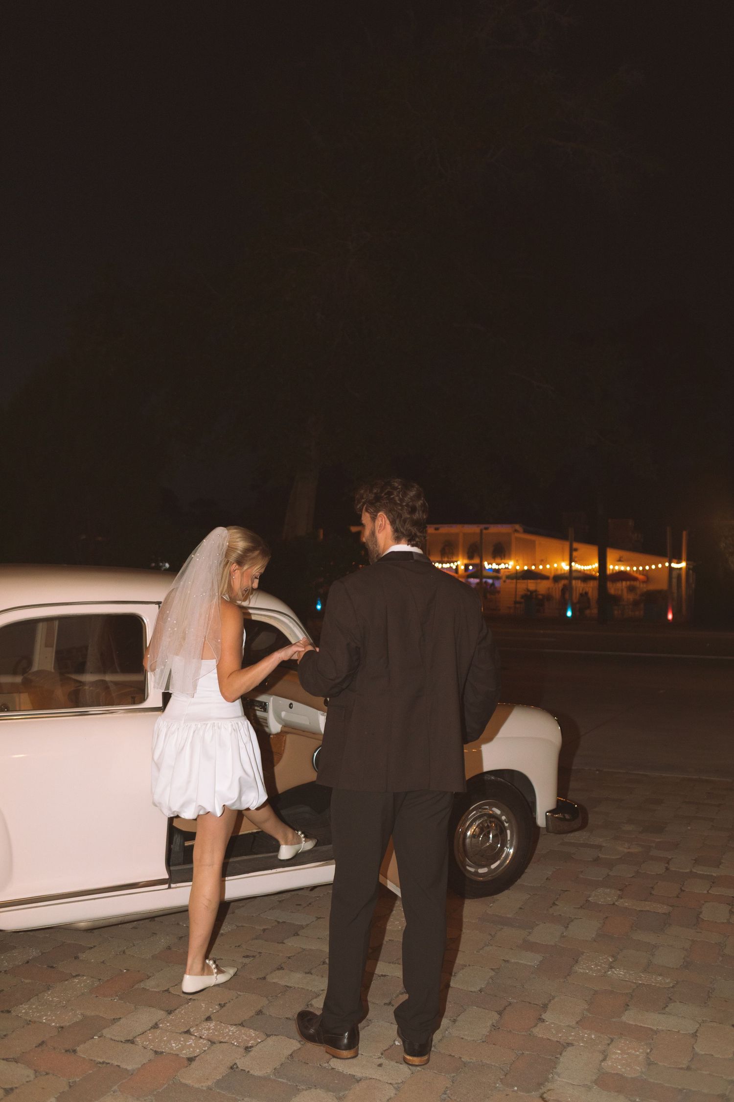 Groom opening vintage white car door for bride at night wedding.