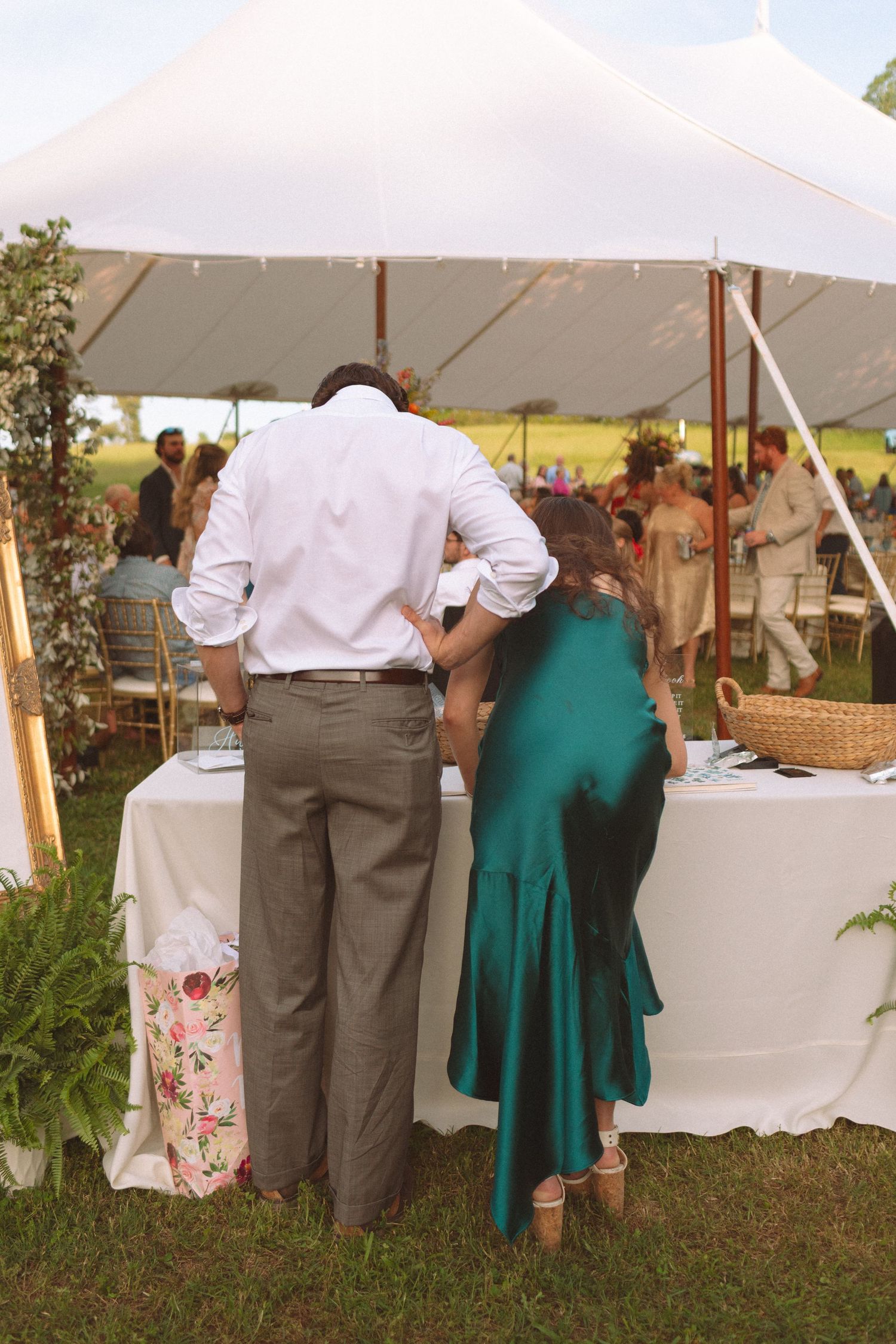 Wedding guests signing in at a white-draped gift table under an outdoor tent at an elegant garden reception.