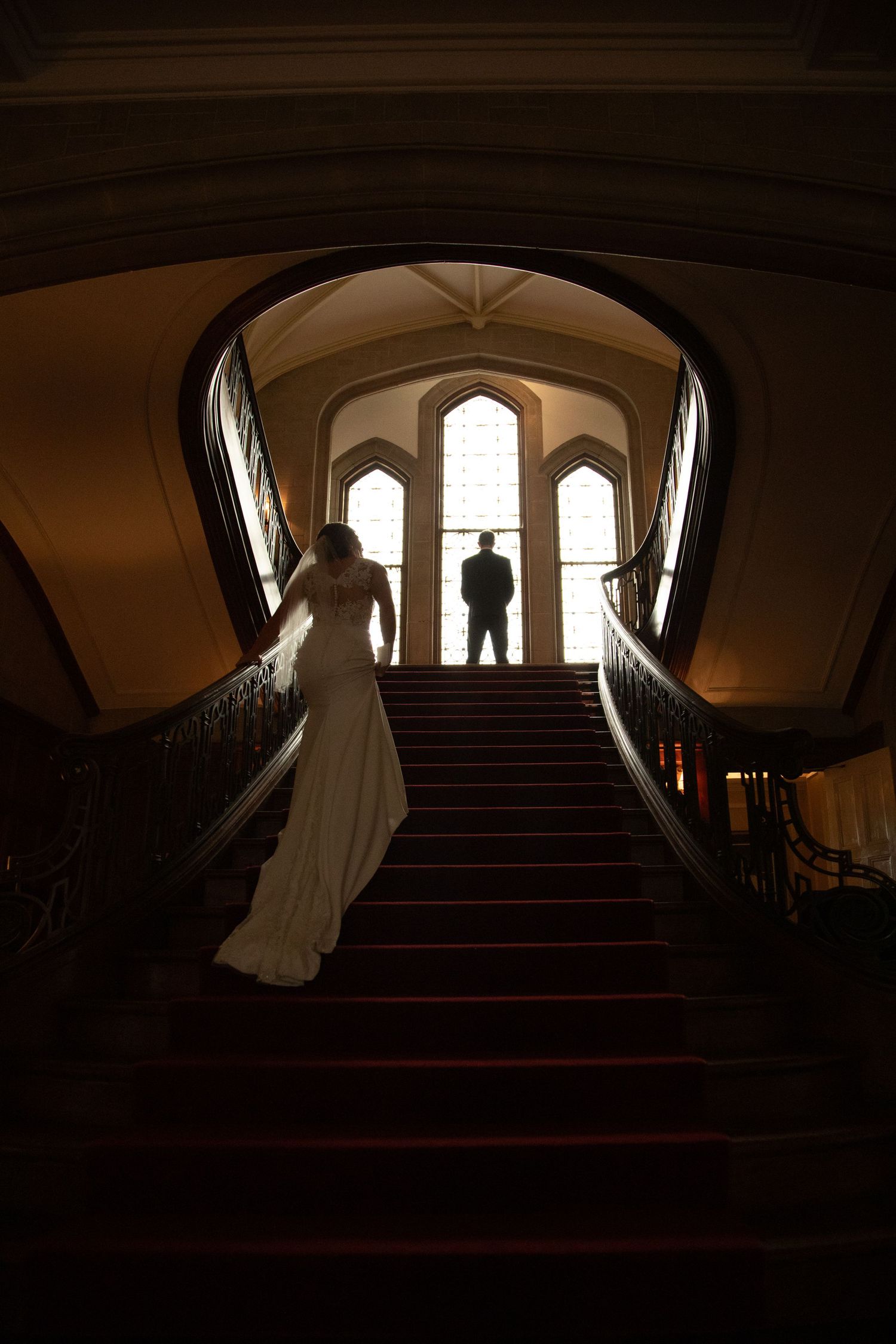Bride ascending grand staircase toward groom silhouetted in arched window of elegant venue.