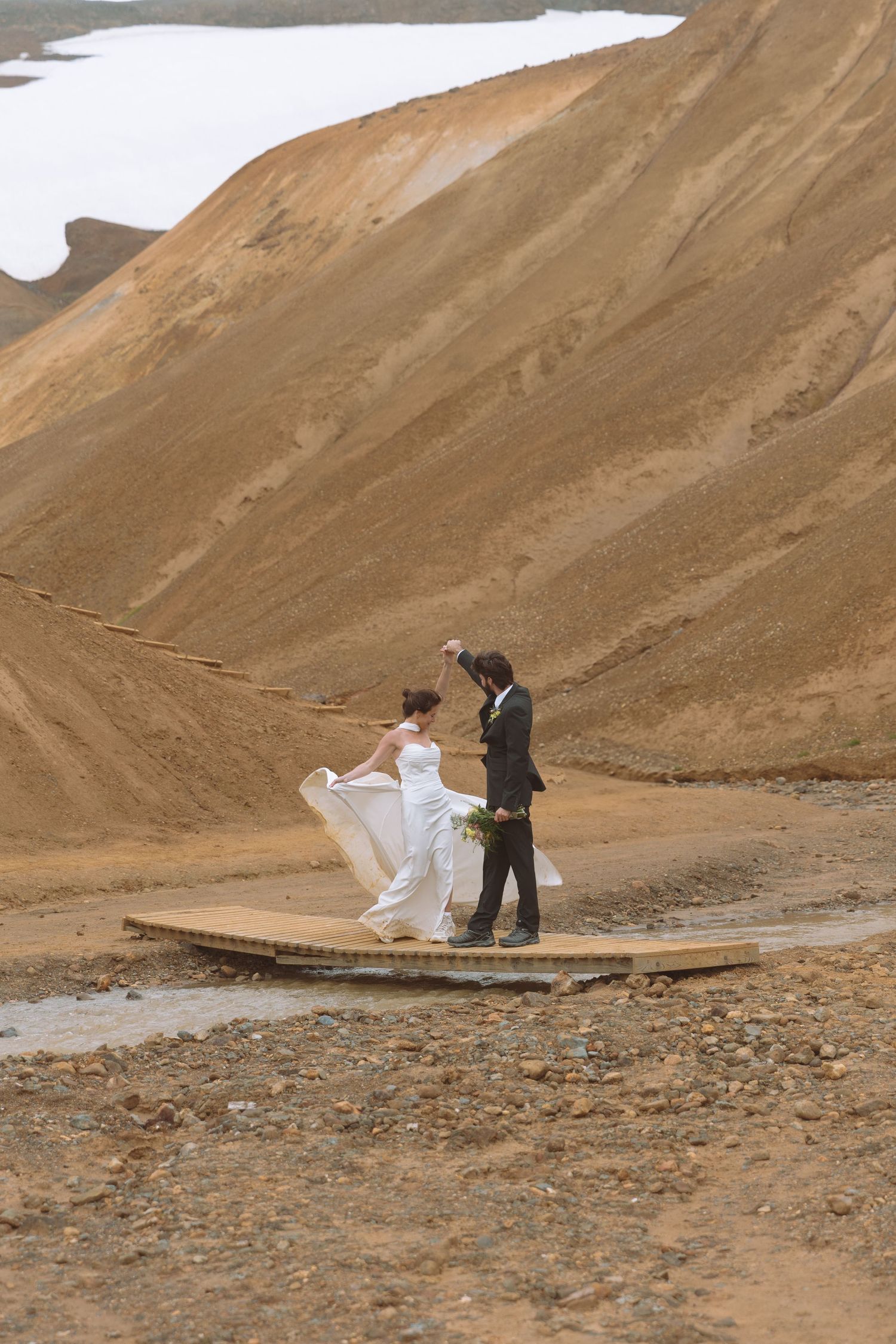 Bride's white gown flows in the wind as couple stands together on a bridge in Iceland's barren landscape.