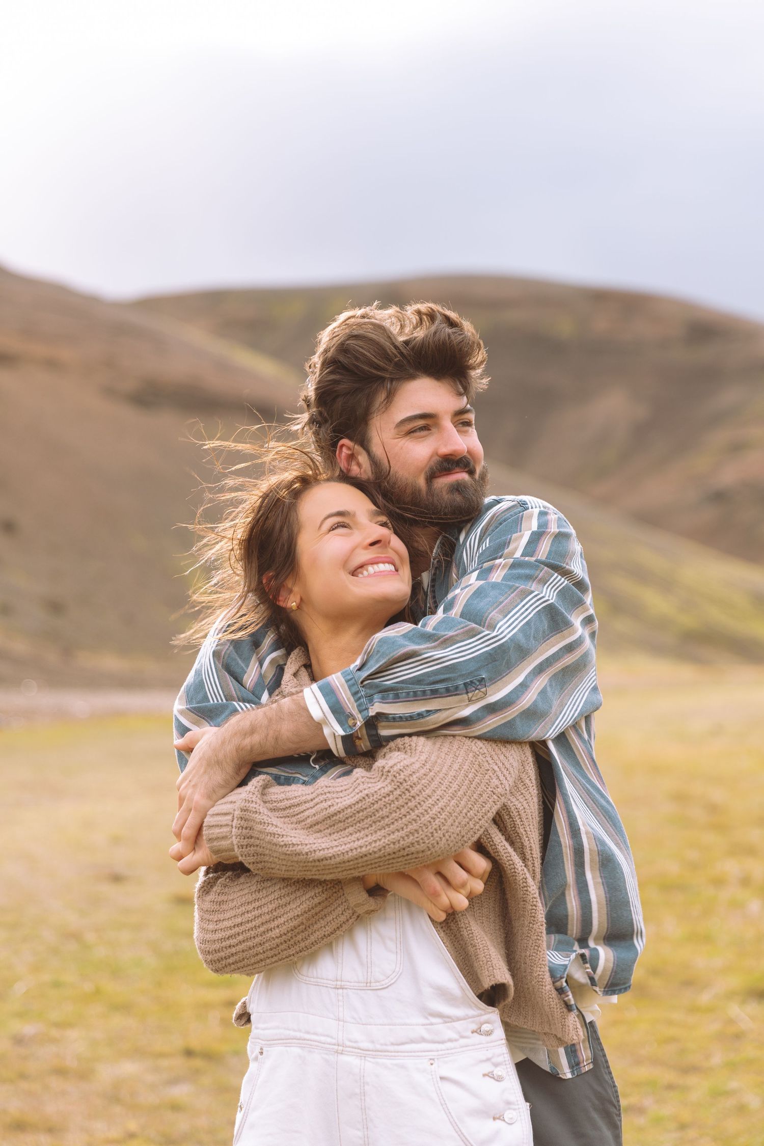 Happy couple hugging outdoors with rolling green hills in the background on a cloudy day.