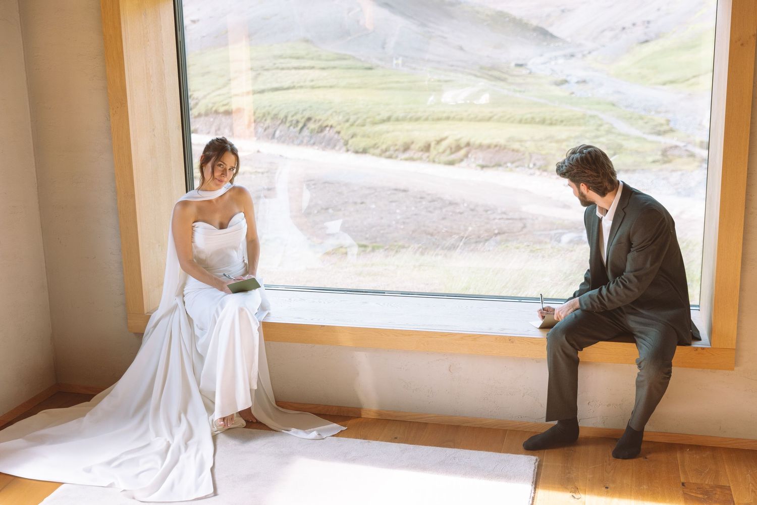A bride in a white gown and groom in dark suit sit separately by a large scenic window.