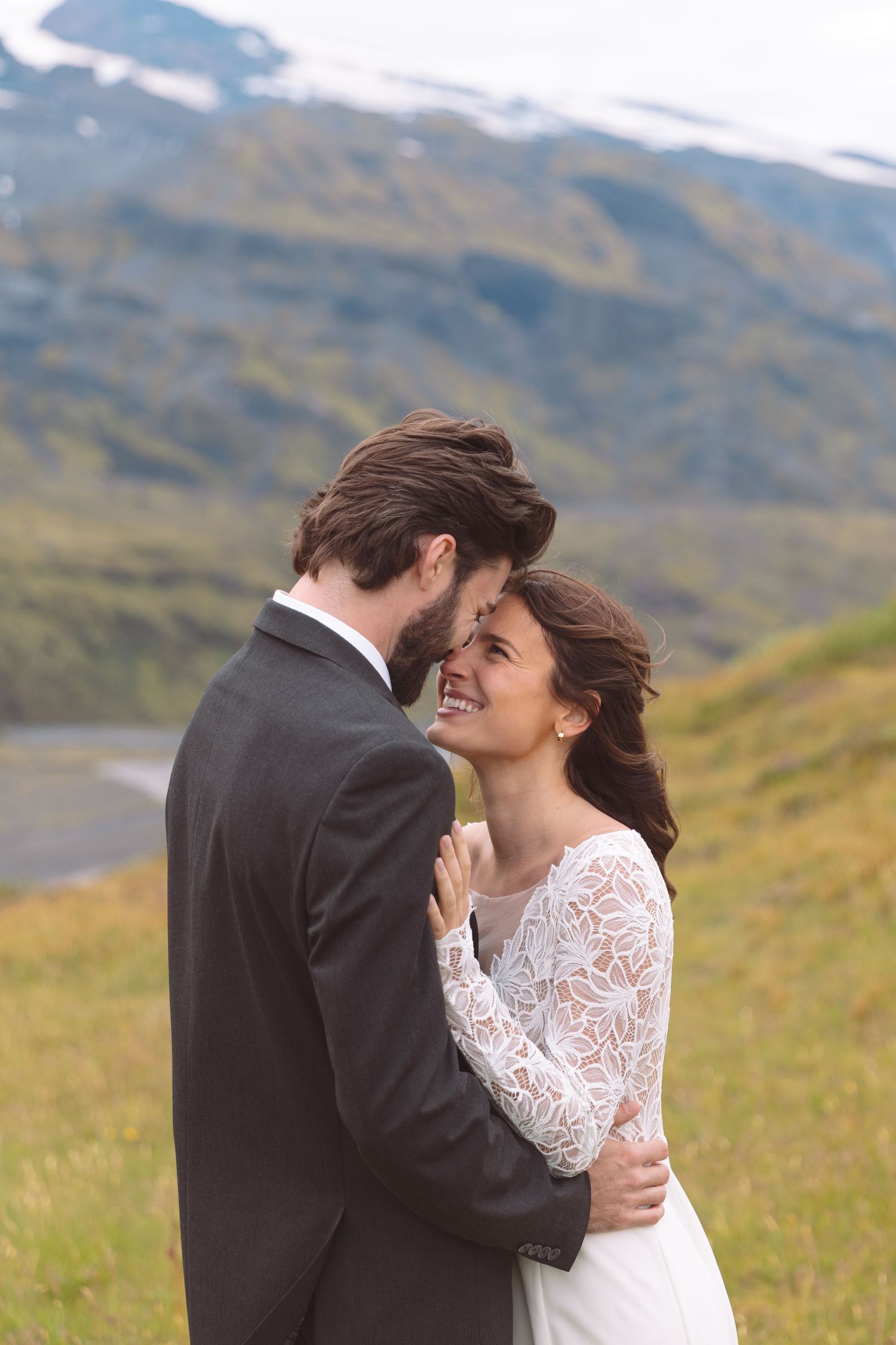 Smiling bride in lace gown embraces groom during outdoor wedding shoot with green mountains backdrop.