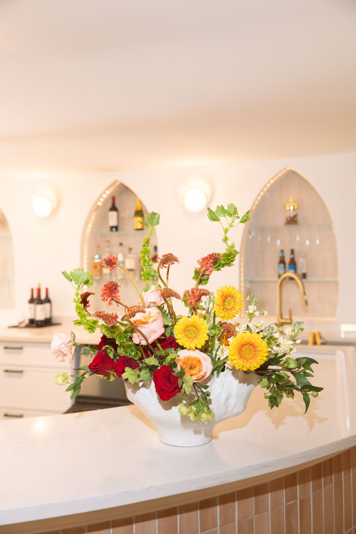 Colorful flower arrangement with yellow, red, and pink blooms in white vase on a bar counter.