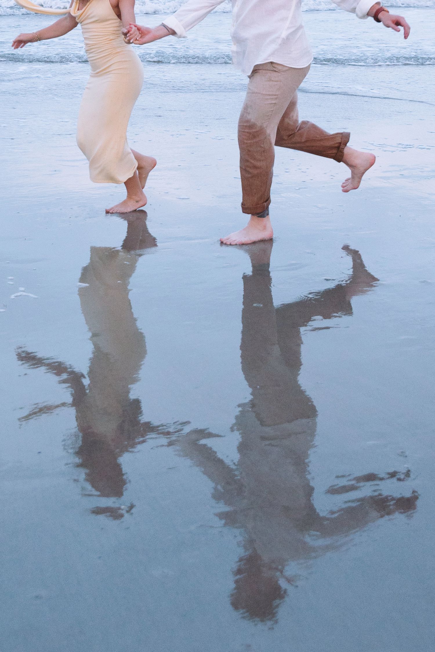 Barefoot couple running on wet beach sand with reflections visible, wearing light casual clothes at the shoreline.