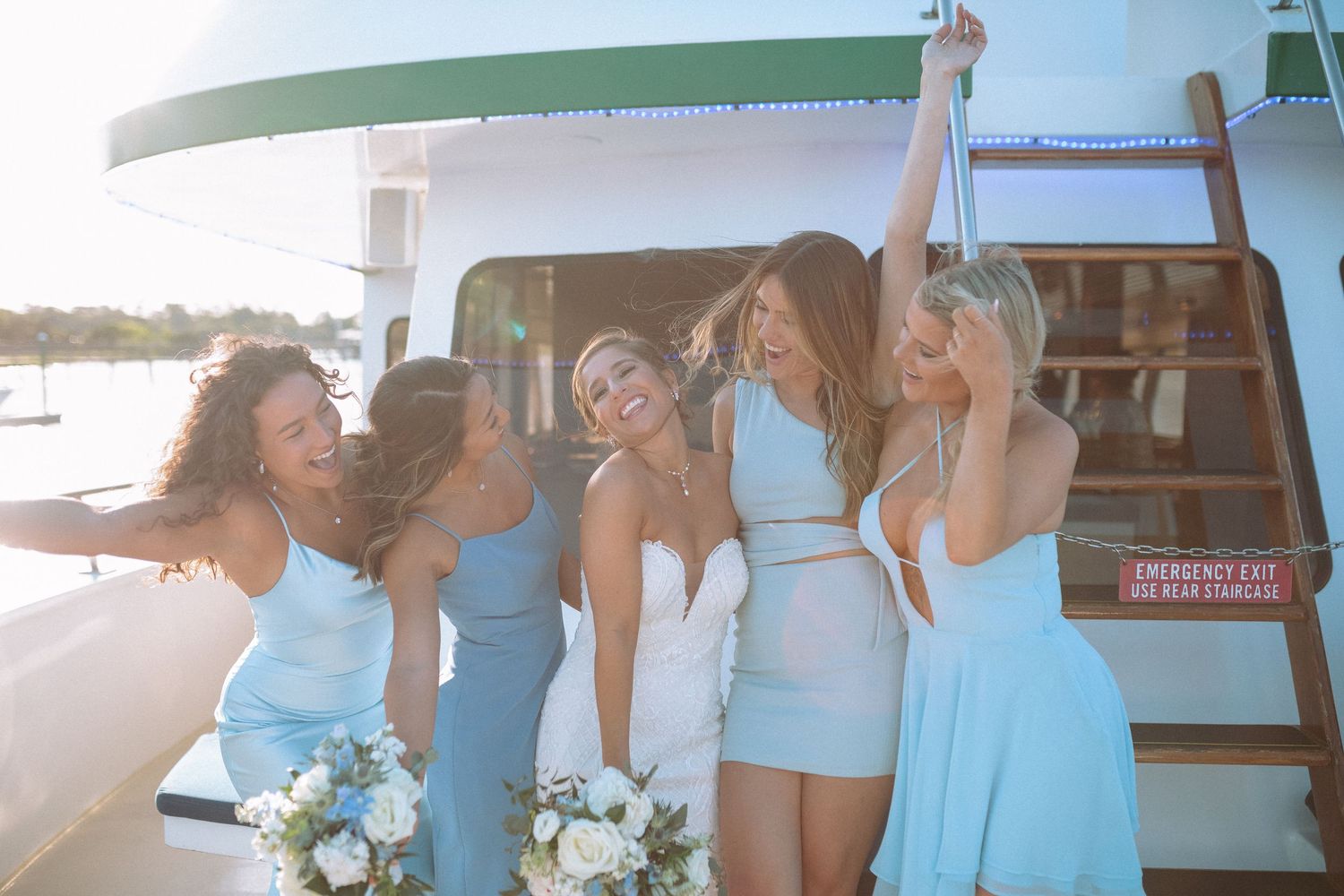 Bride and bridesmaids in blue dresses laughing together on a sunny boat deck.