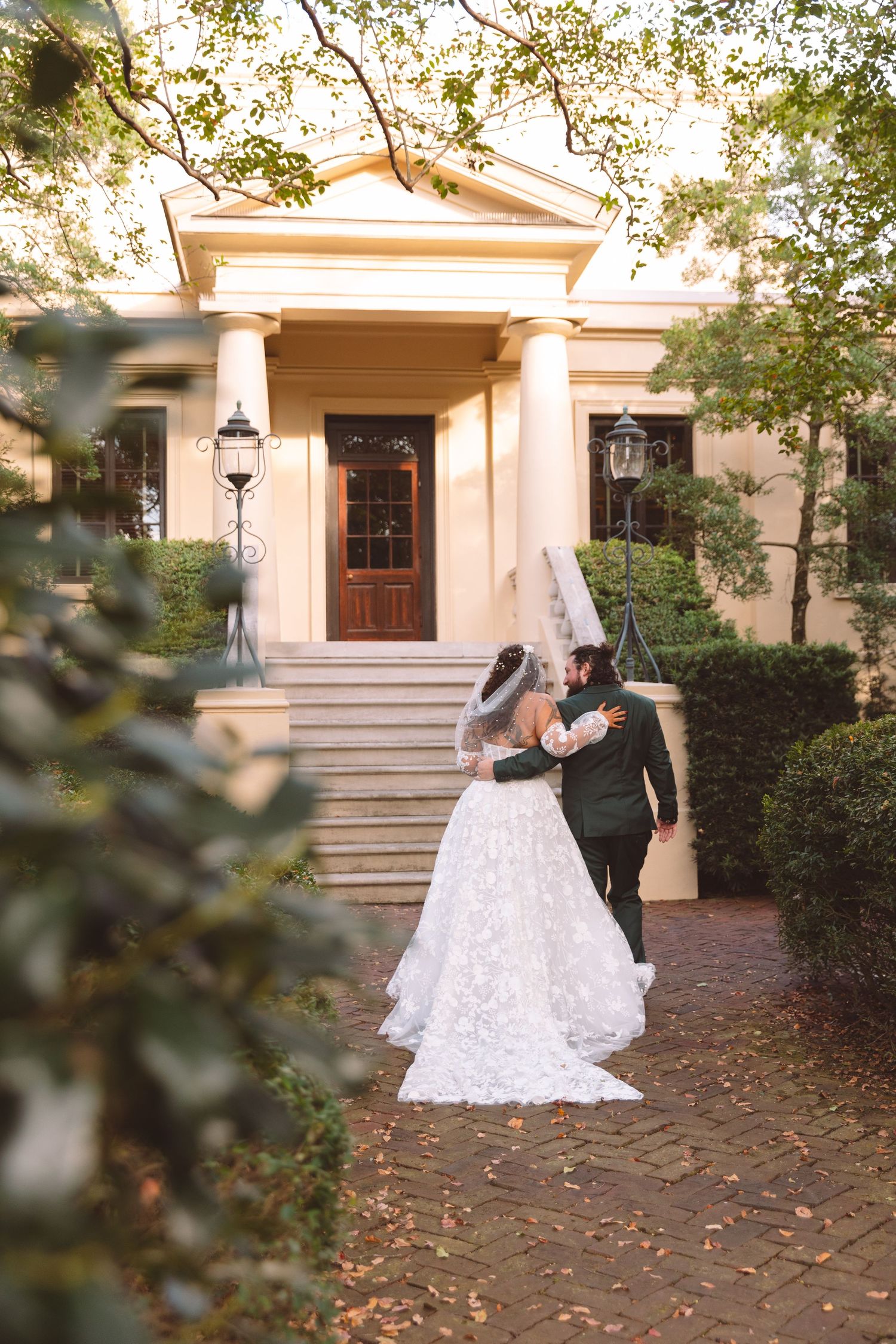 Bride and groom embracing on brick pathway in front of a grand Southern estate venue.