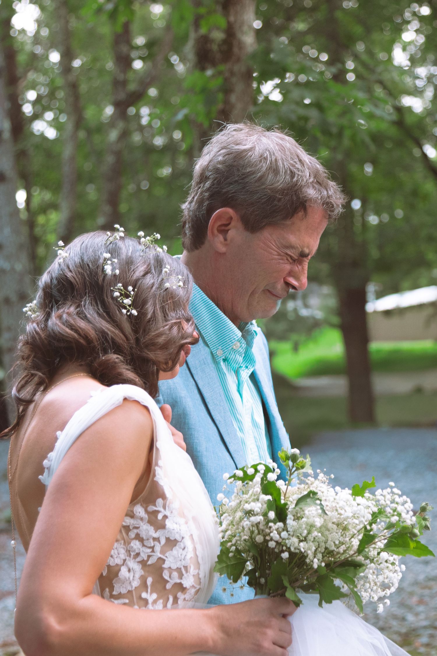Bride with floral hair accessories holding white bouquet standing with father in turquoise suit outdoors.