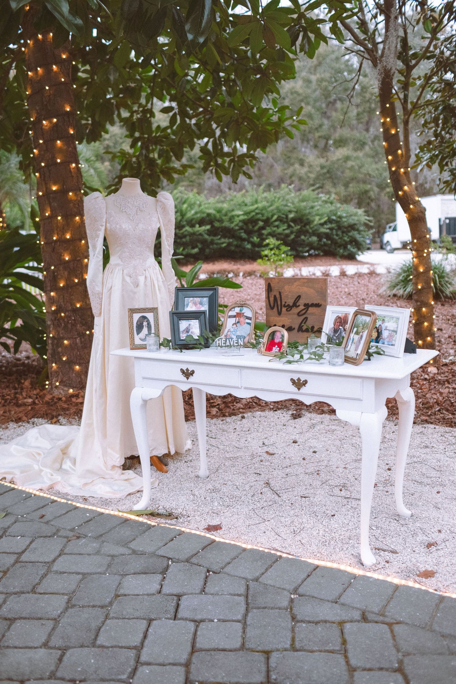 Elegant outdoor wedding display with white dress on mannequin, fairy lights on trees, and framed photos on white vintage table.