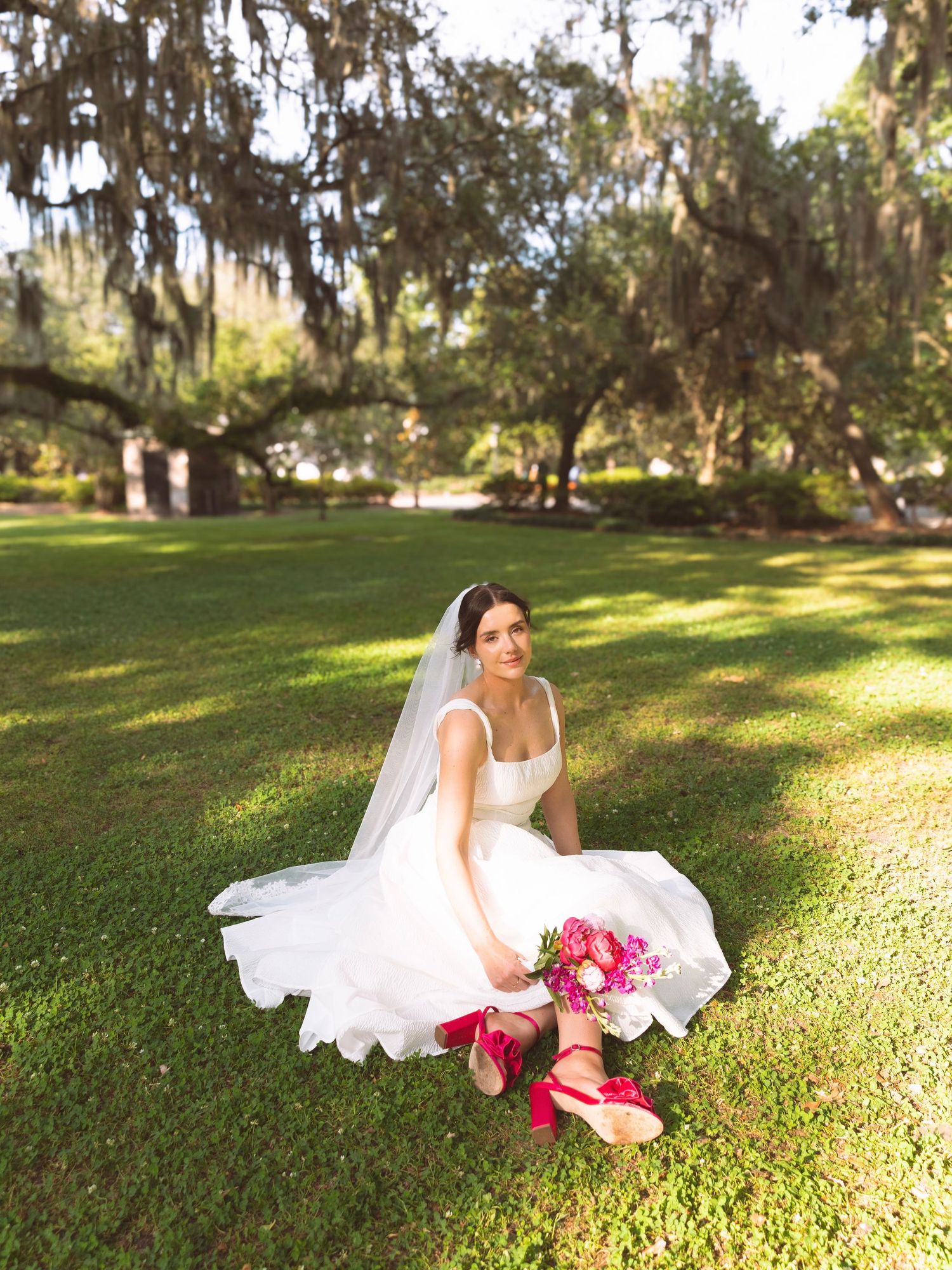 Bride in white gown and veil sitting on grass adjusting pink heels while holding a pink bouquet.