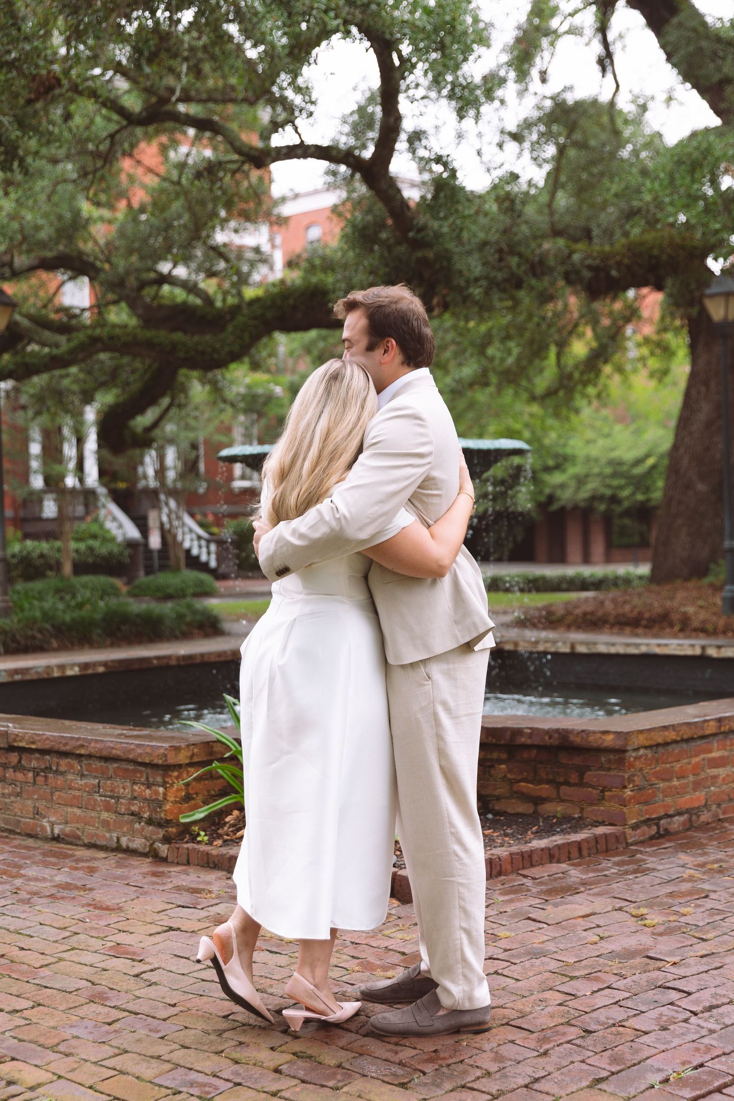 Newlyweds embrace in romantic hug beside brick fountain under large oak tree after outdoor ceremony.