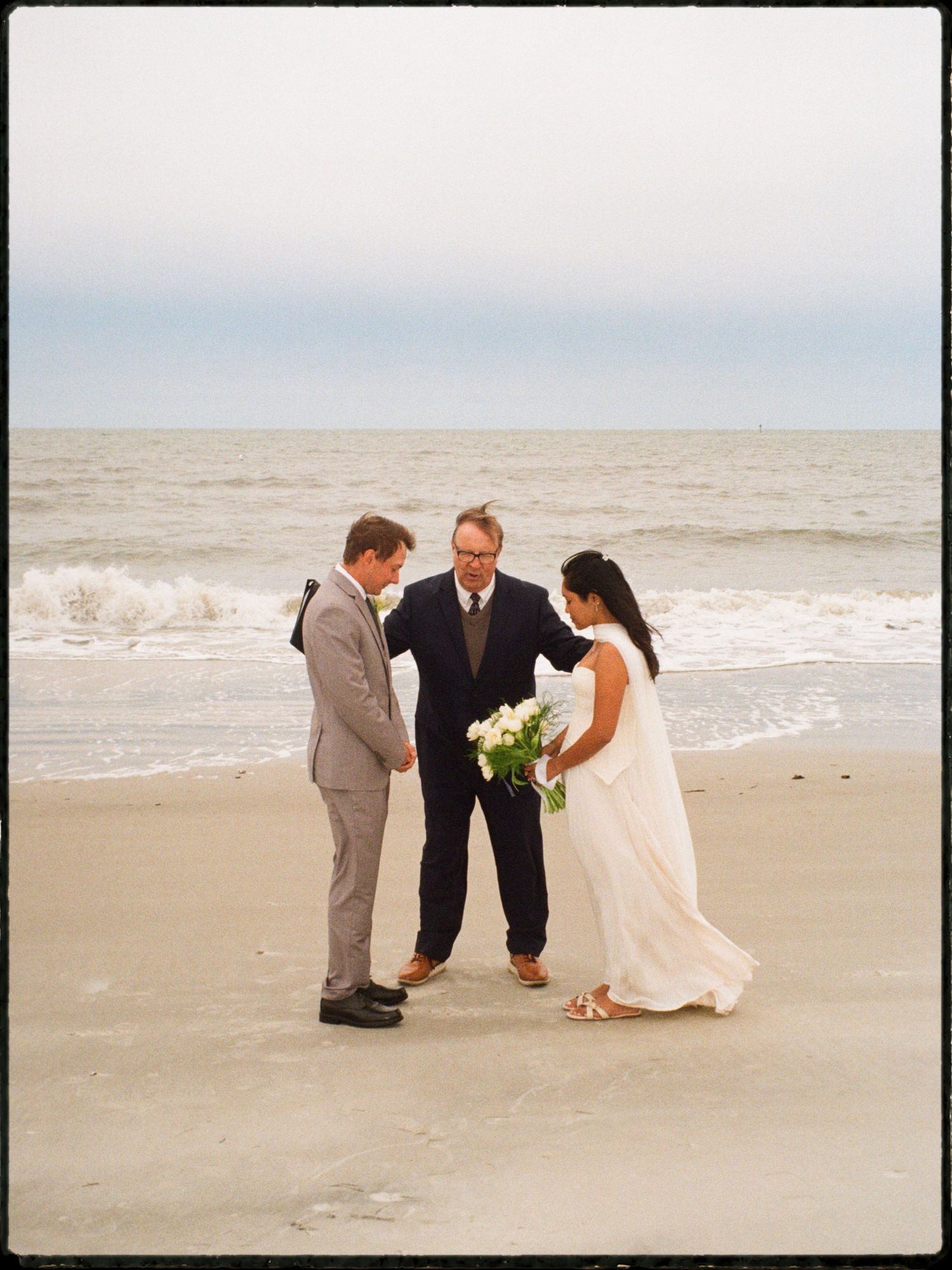 Bride and groom exchange vows on a beach with an officiant, bride holding white flowers, ocean waves in background.