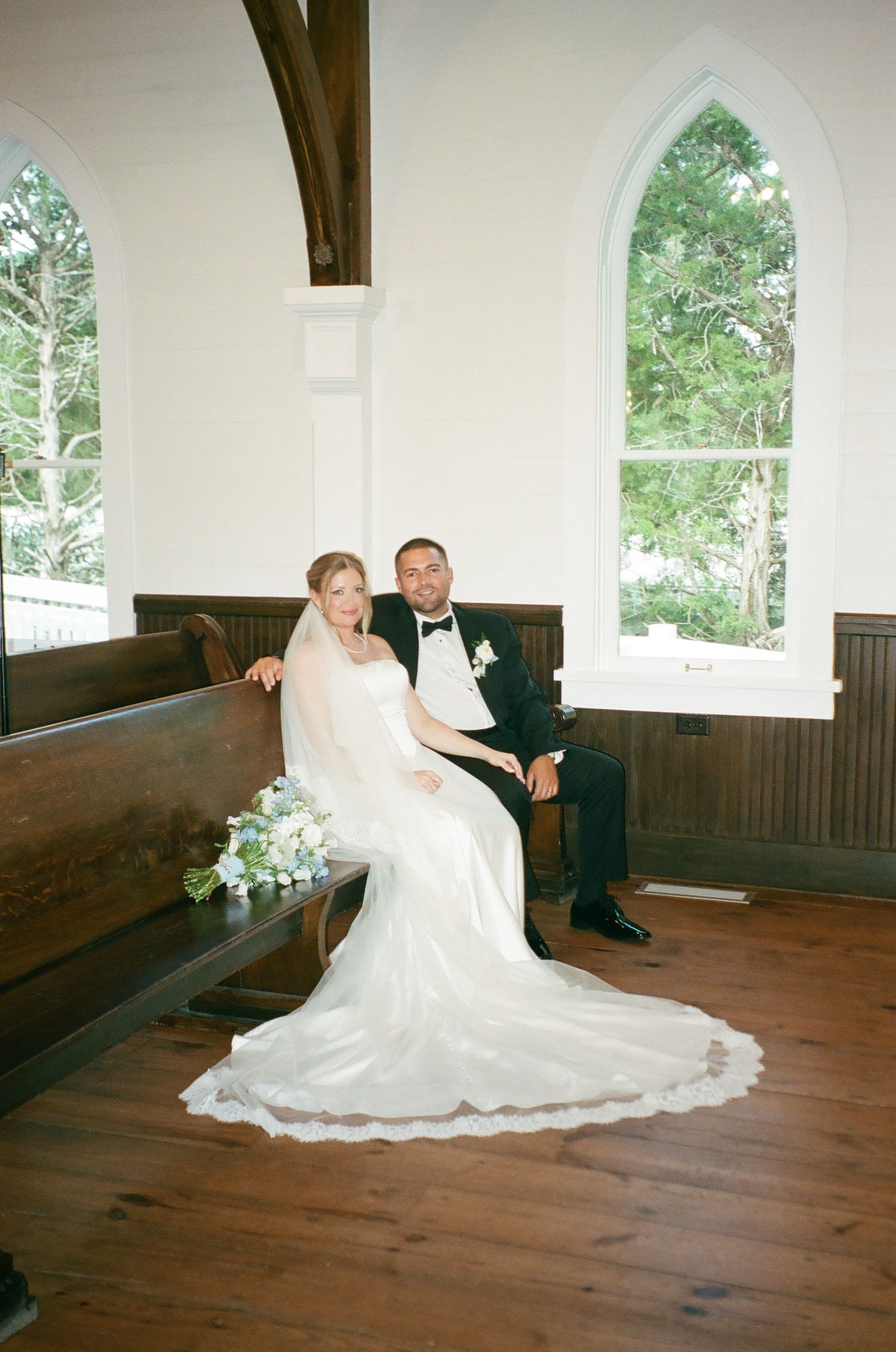 Bride in white gown and groom in tuxedo sit together on church pew with floral bouquet in rustic chapel.