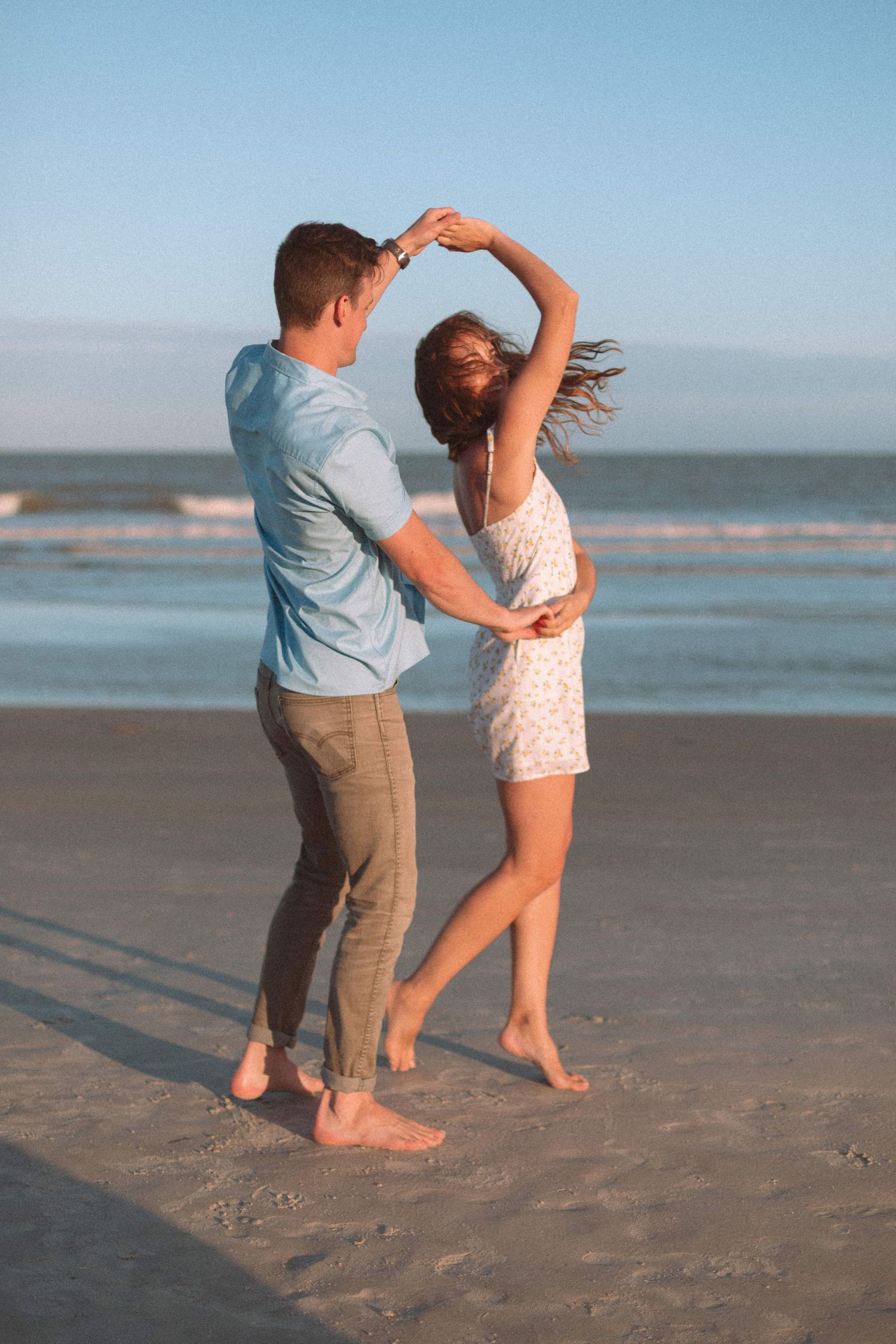 Couple dancing barefoot on a sandy beach at sunset, man spinning woman in a white floral dress.