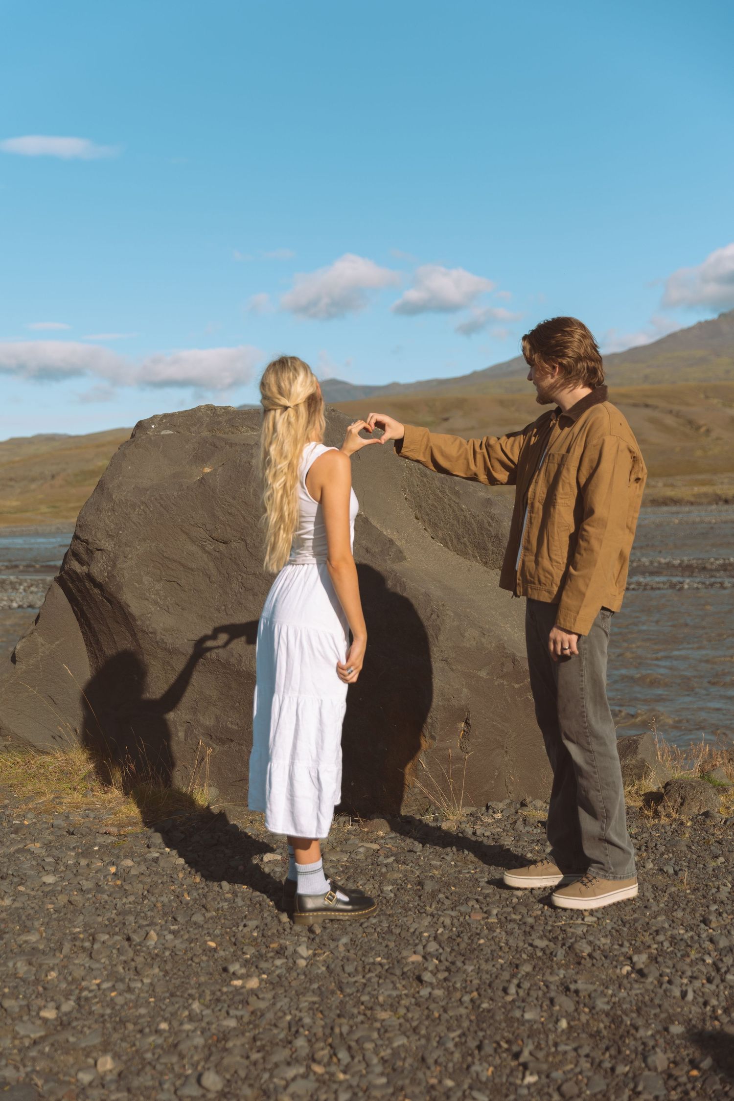Couple shares a tender heart gesture beside a large boulder in a rugged outdoor setting.