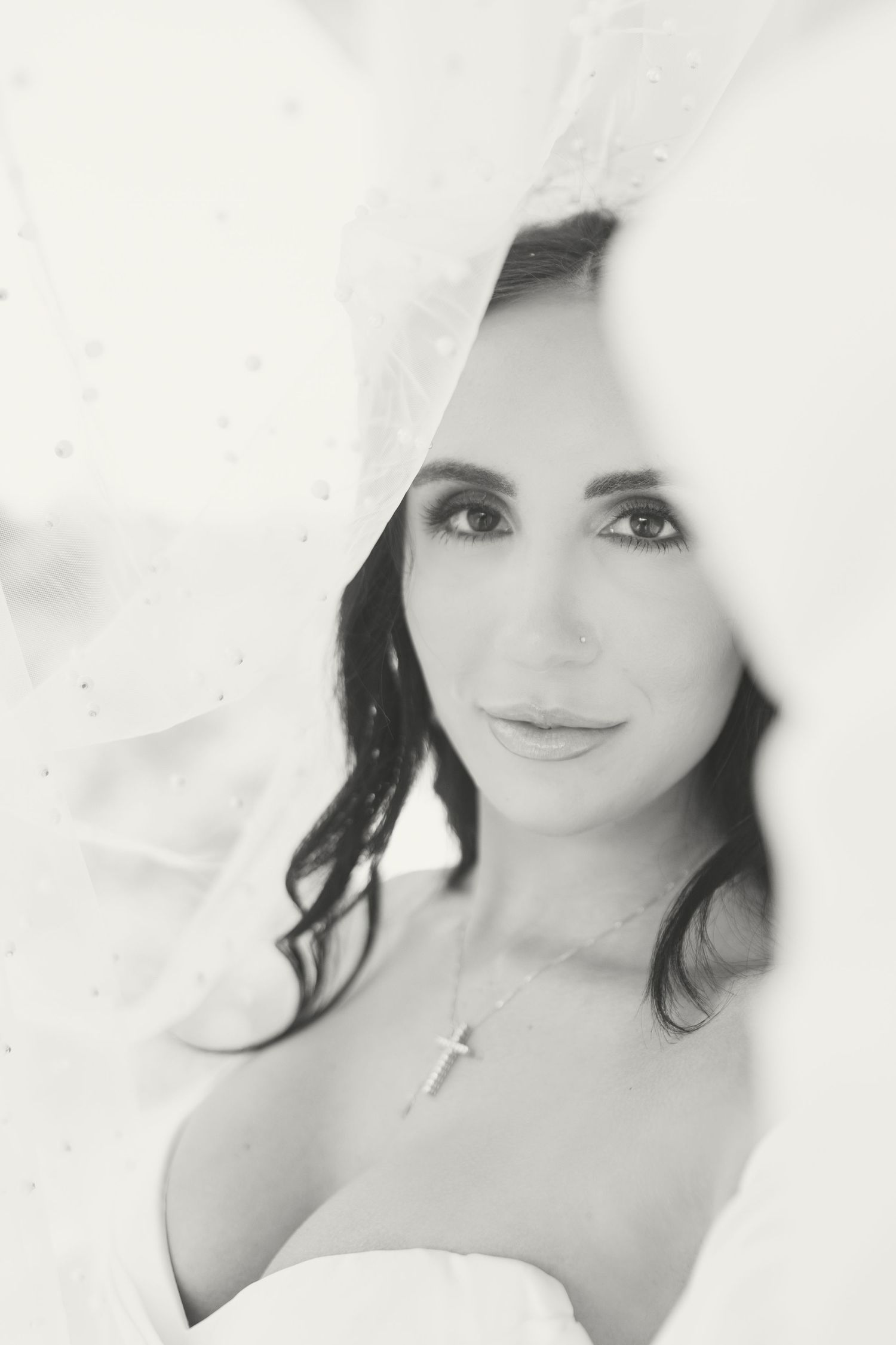 Black and white bridal portrait of a dark-haired bride wearing a cross necklace, framed by a flowing white veil.