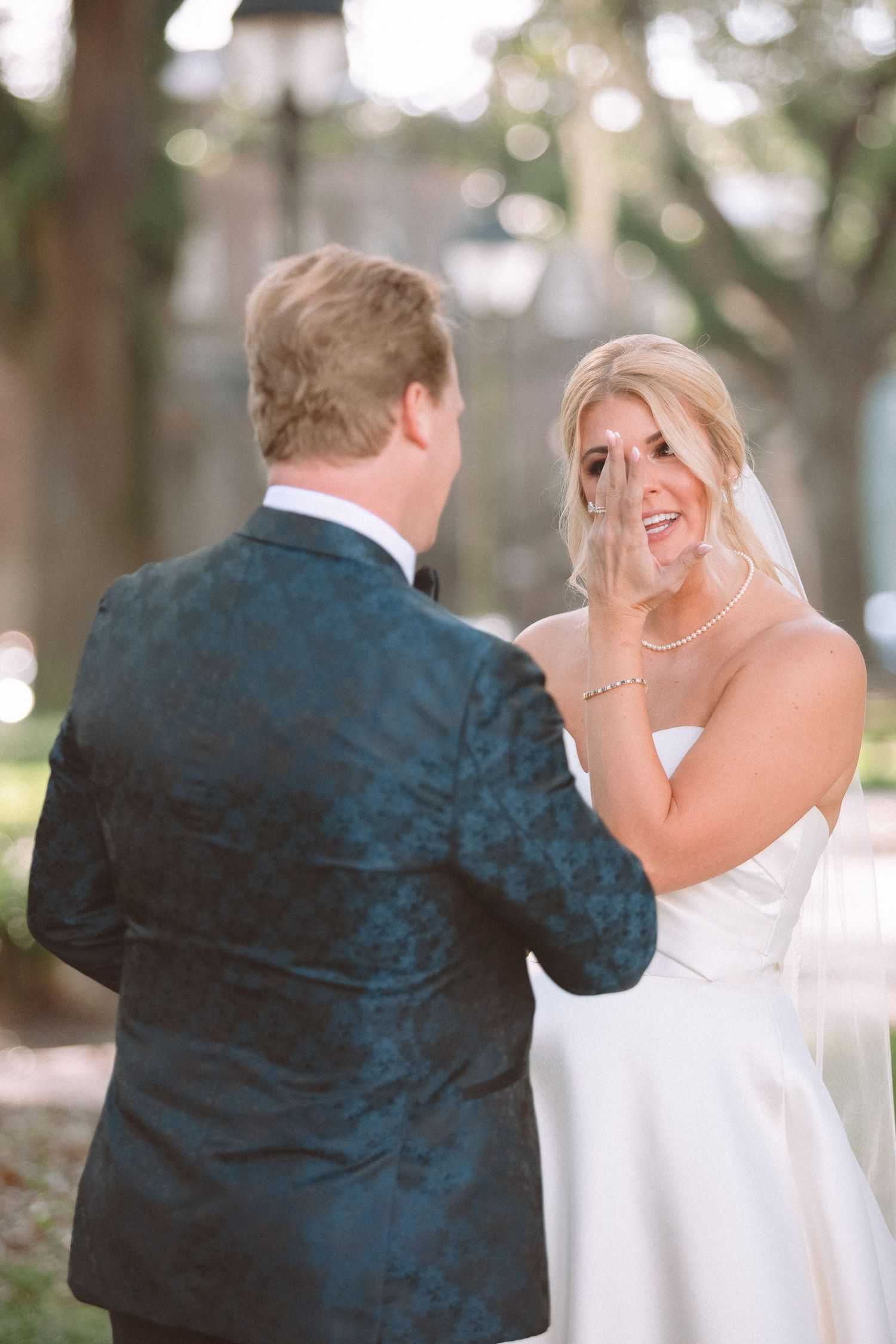 Emotional bride wiping tears during first look moment with groom outdoors.