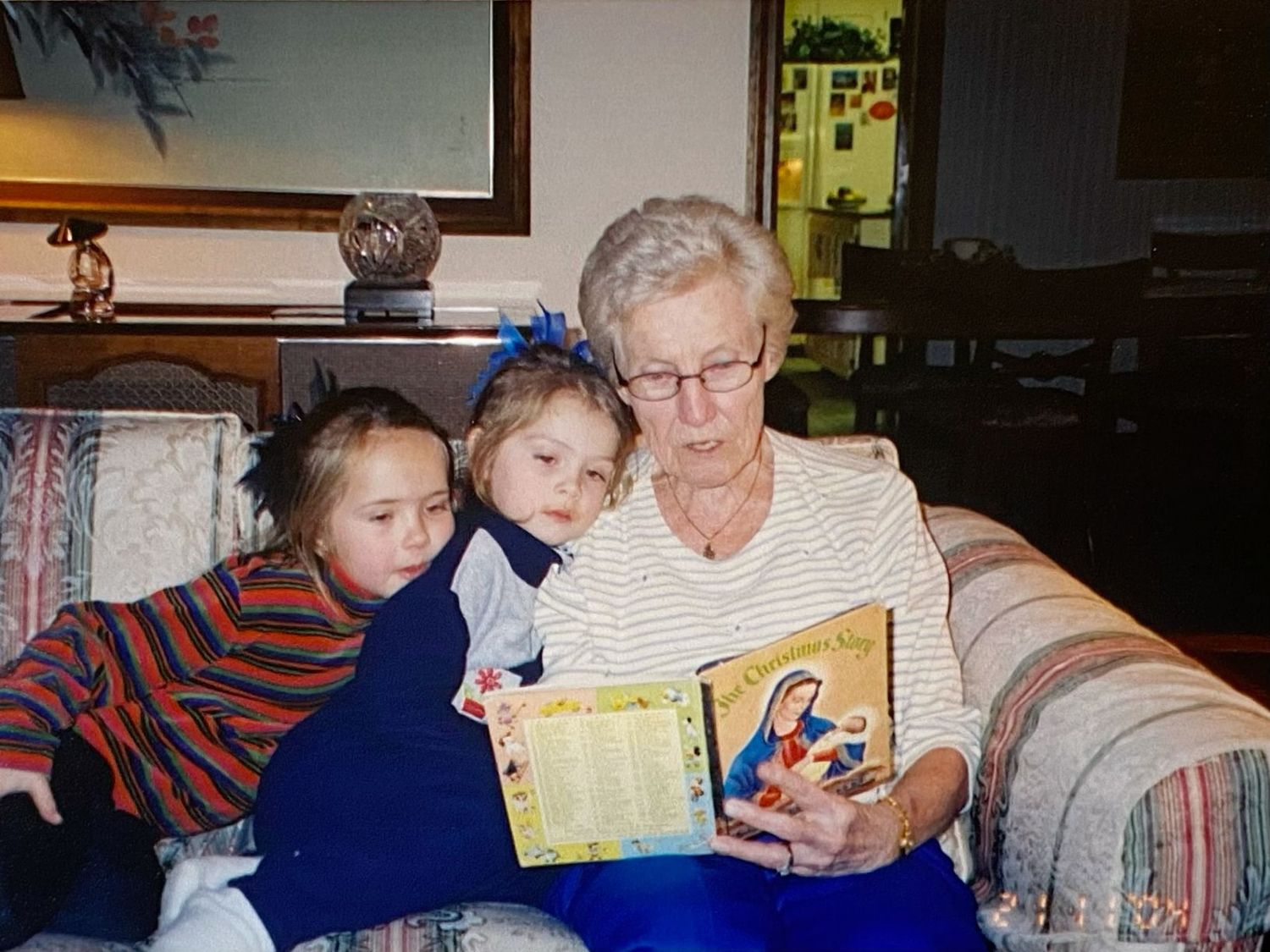Elderly grandmother reads a colorful religious book to two young girls on a floral sofa.