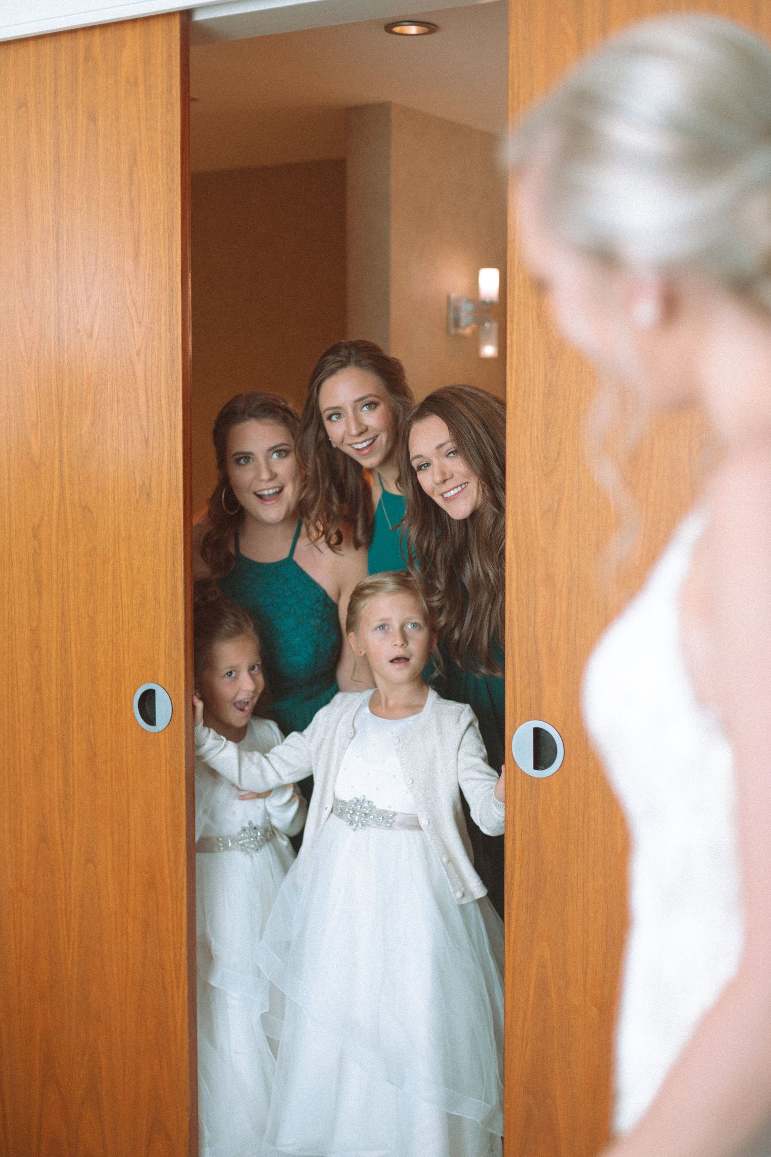 Bridesmaids in teal dresses and flower girls react with awe at bride's first look reveal.