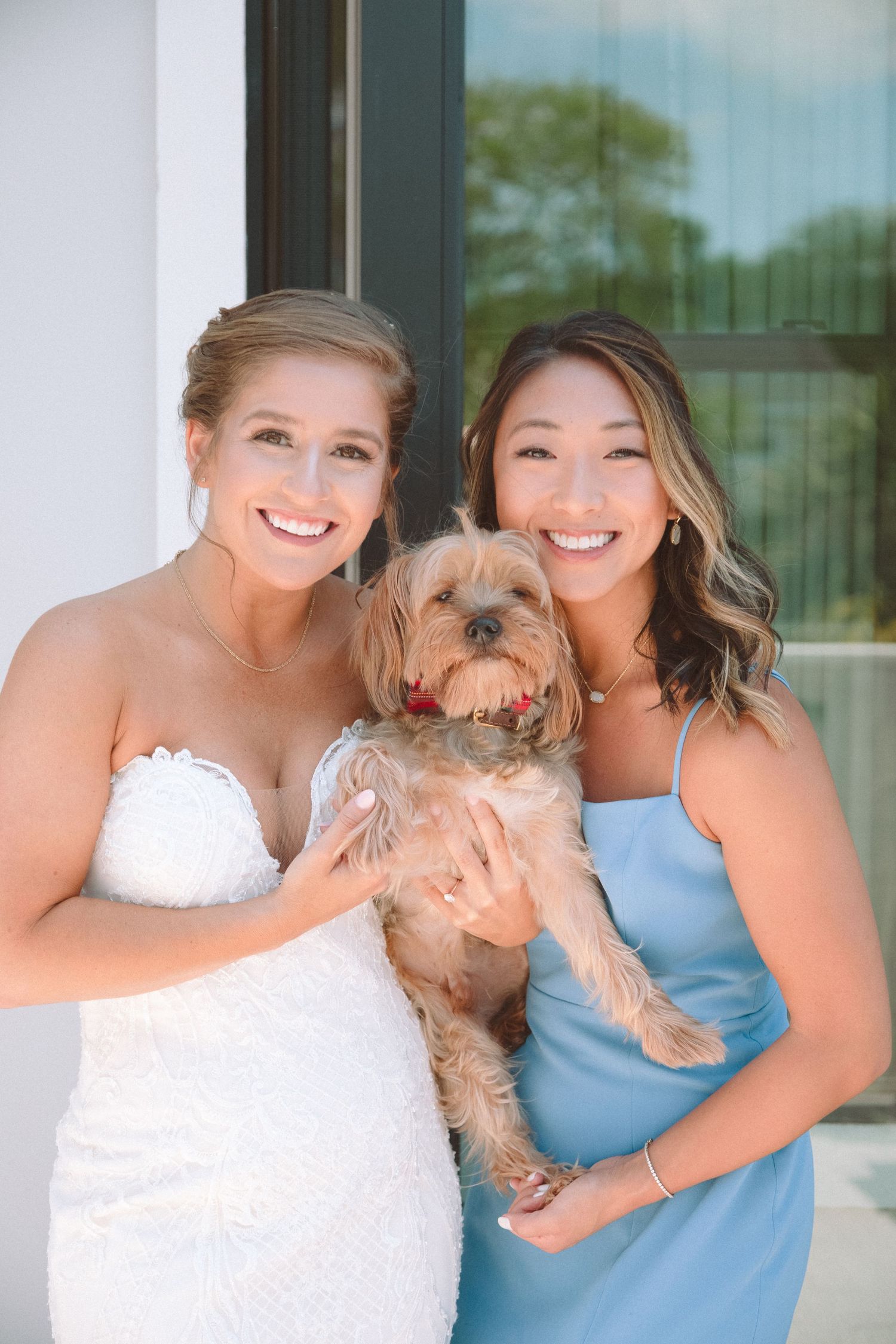 Smiling bride in white strapless gown and bridesmaid in blue dress posing with a small fluffy dog.