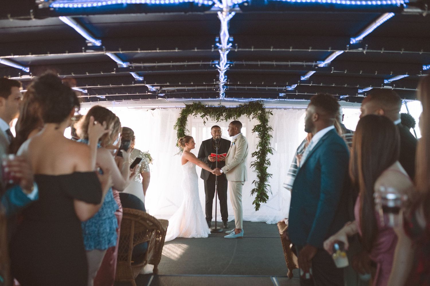Bride and groom exchanging vows under a greenery arch at an intimate indoor ceremony.