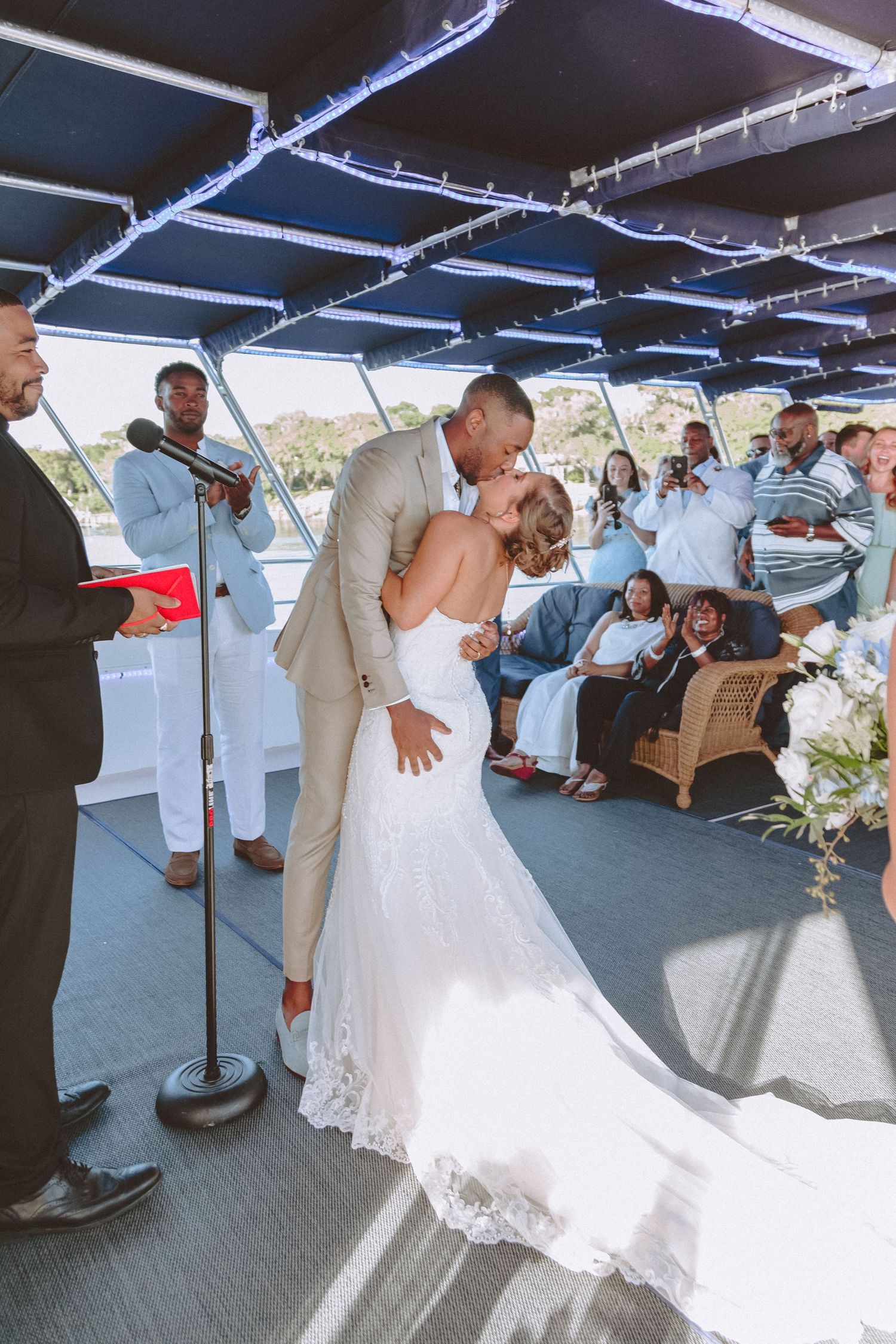 Groom dips bride for kiss at altar as wedding guests cheer during ceremony on boat.