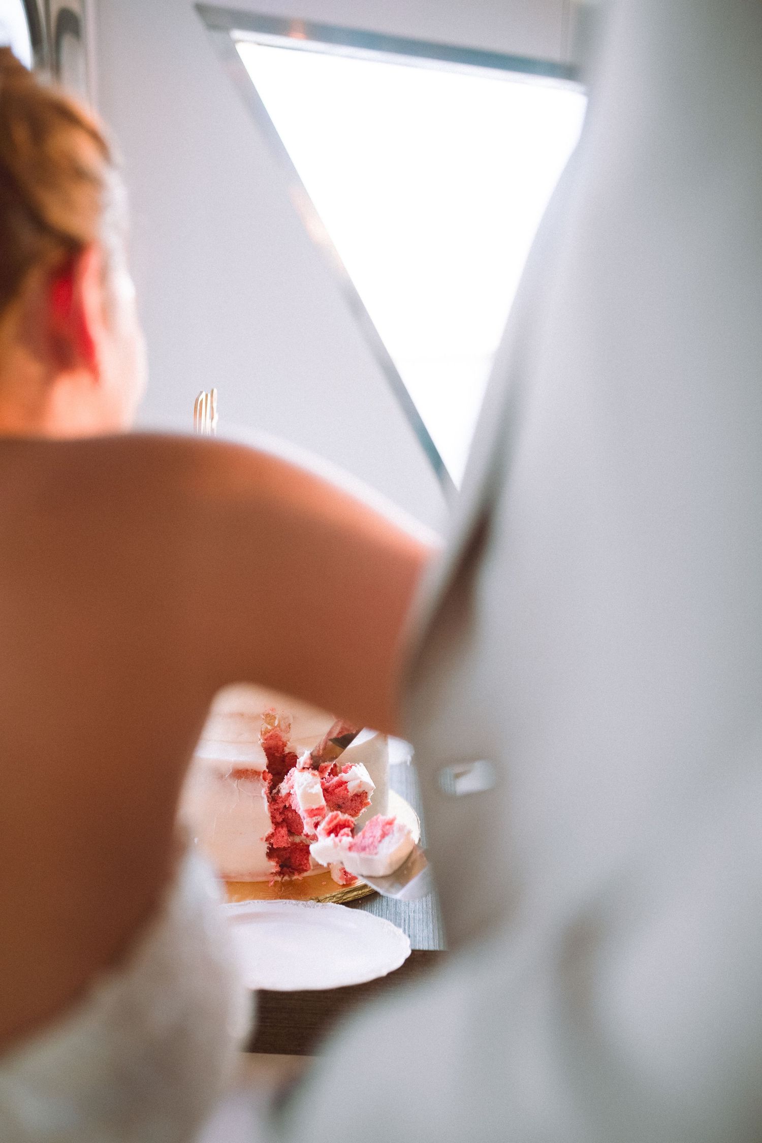 Bride viewed from behind arranging pink floral bouquet near a bright window on wedding day.