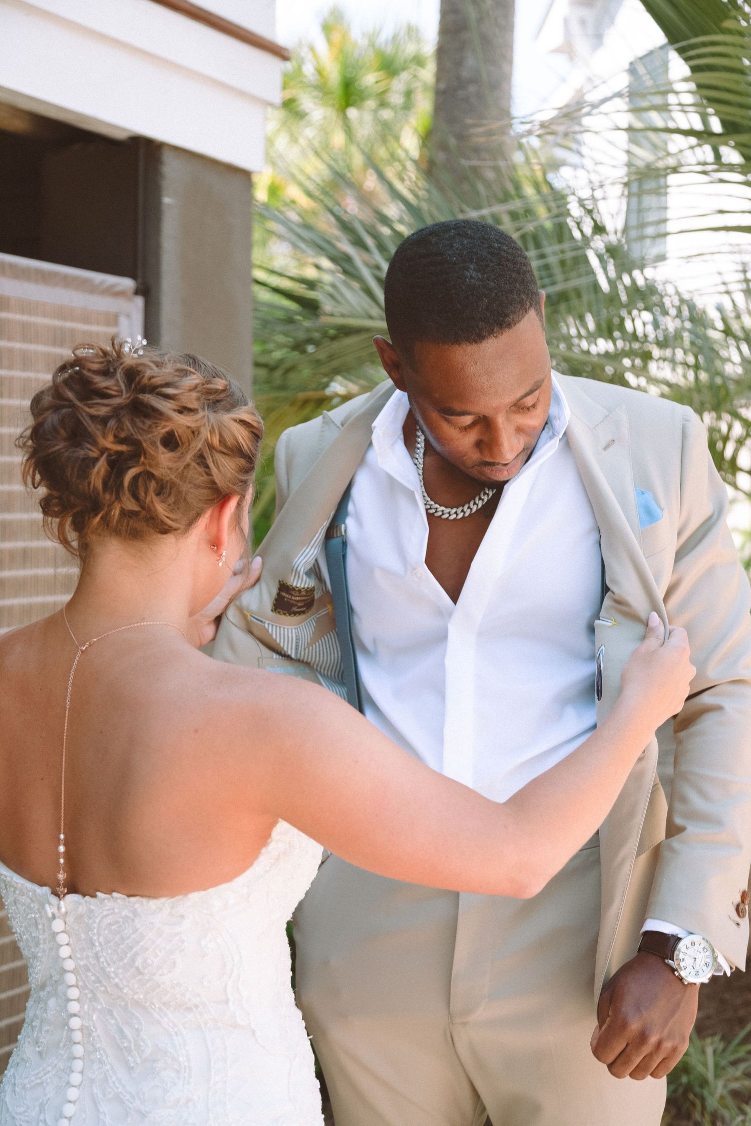 Bride helps straighten groom's tan suit jacket during intimate wedding first look near tropical palms.