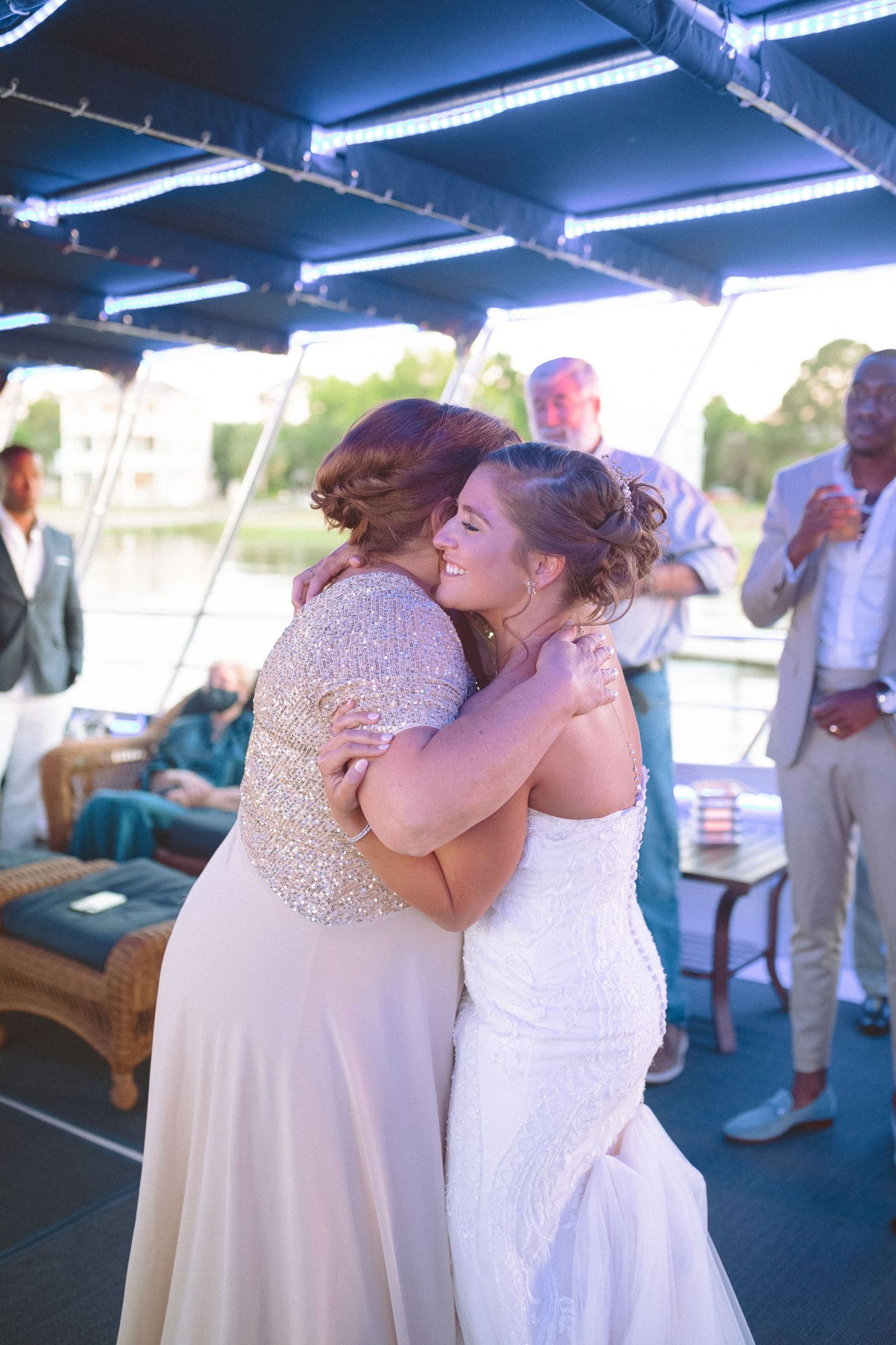 Bride hugs a woman in a gold sequin dress during wedding reception on a boat.