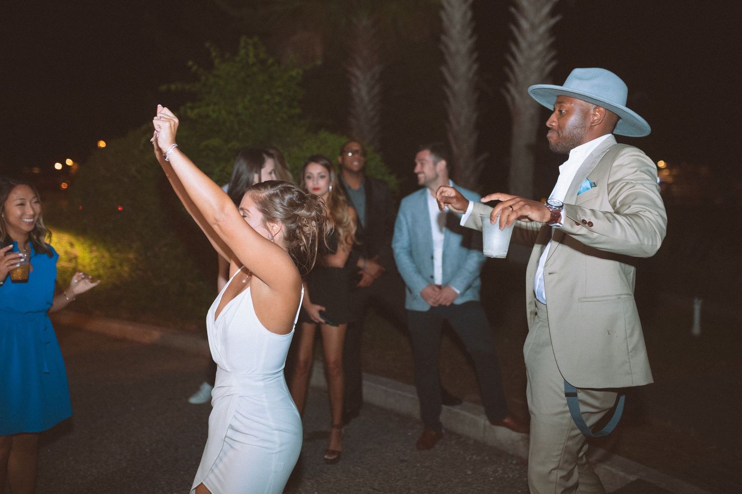 Bride in white dress and groom in tan hat dance outdoors at night with wedding guests watching.