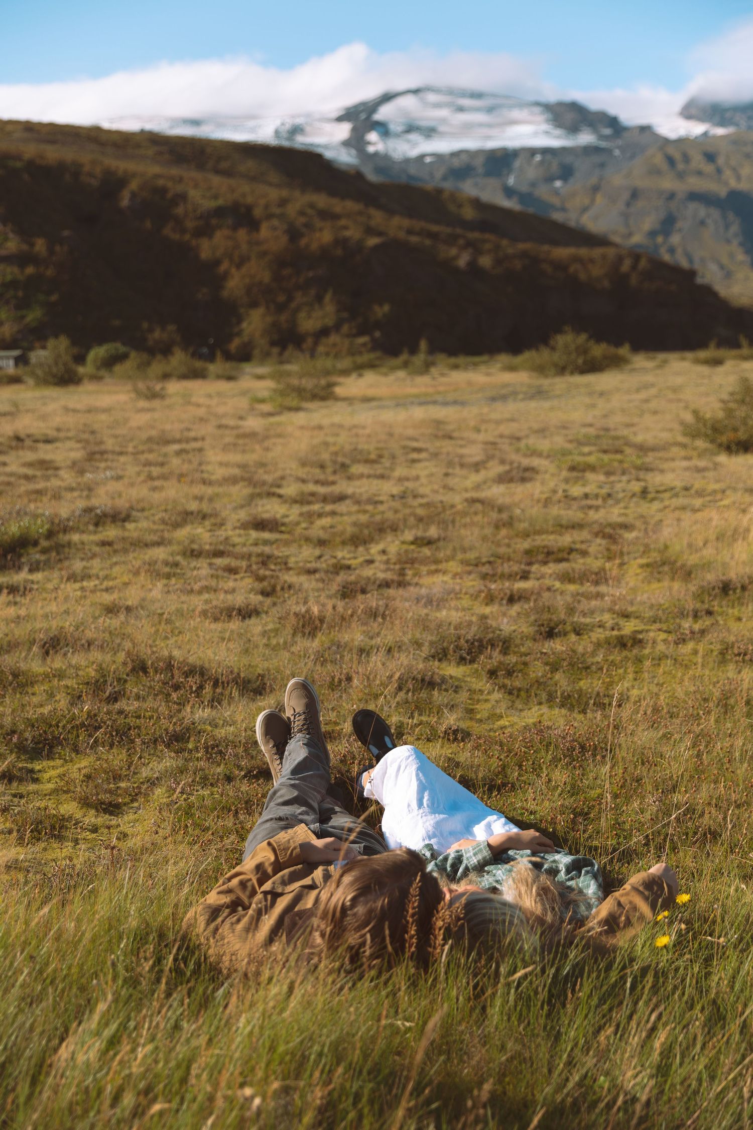 Couple lying in a grassy meadow with snow-capped mountains in the background on a sunny day.