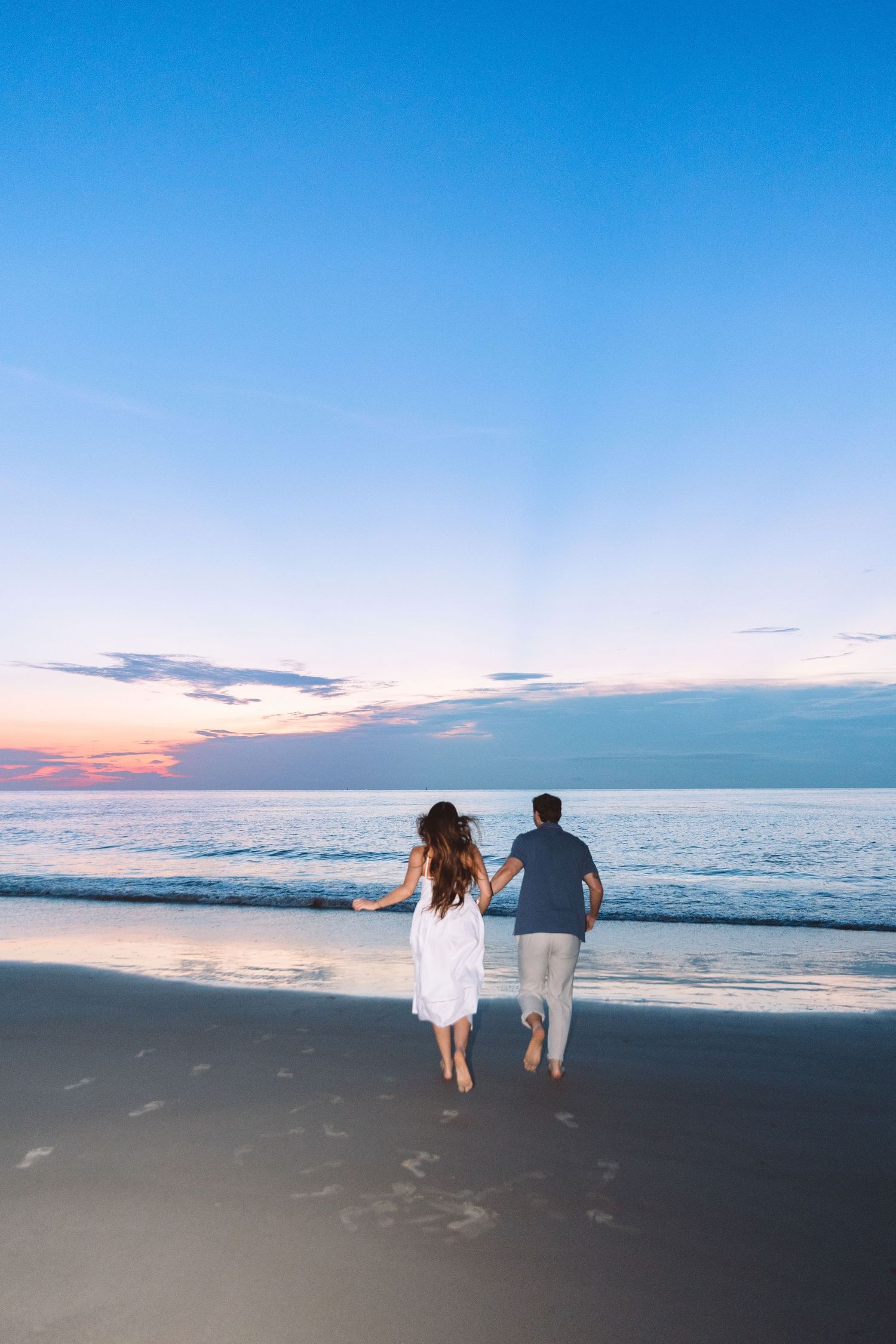 Couple holding hands running toward ocean waves on a sandy beach during a colorful sunset.