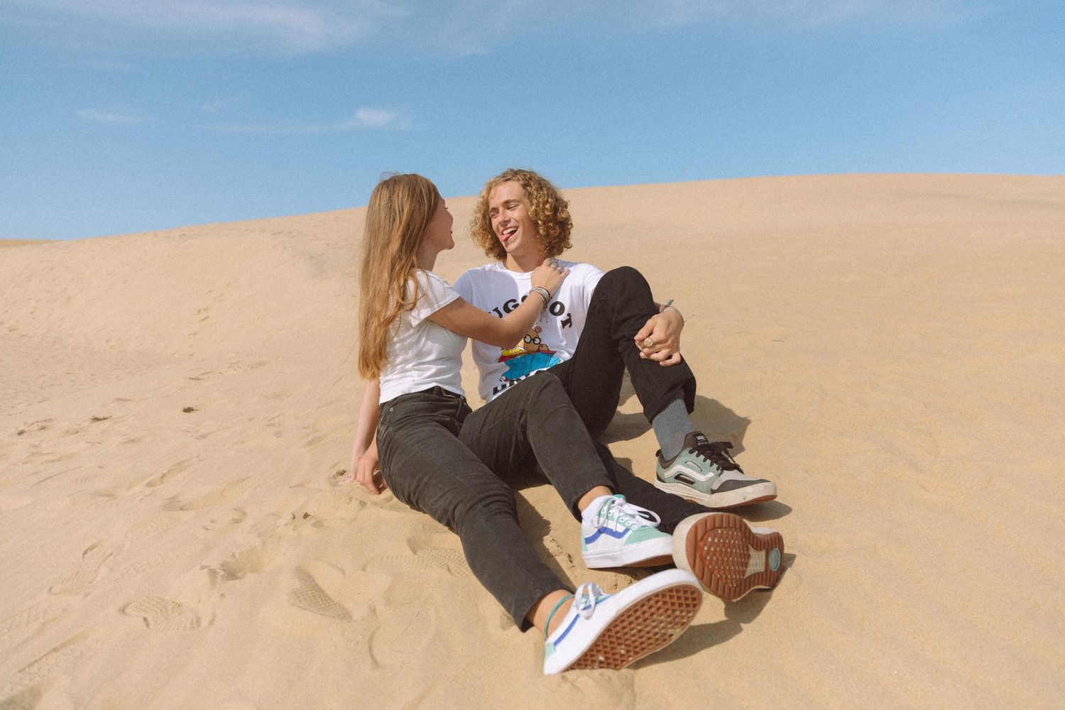 Two friends sitting together laughing on sandy desert dunes.