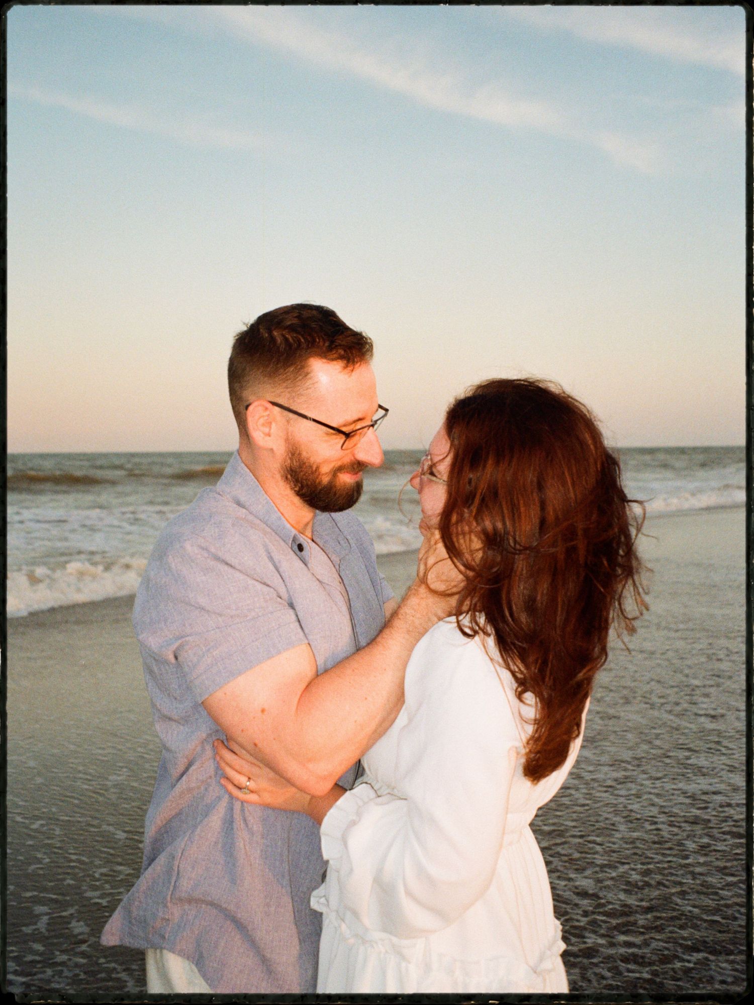 Bearded man gently touching redhead womans face during romantic beach sunset moment on film photo.