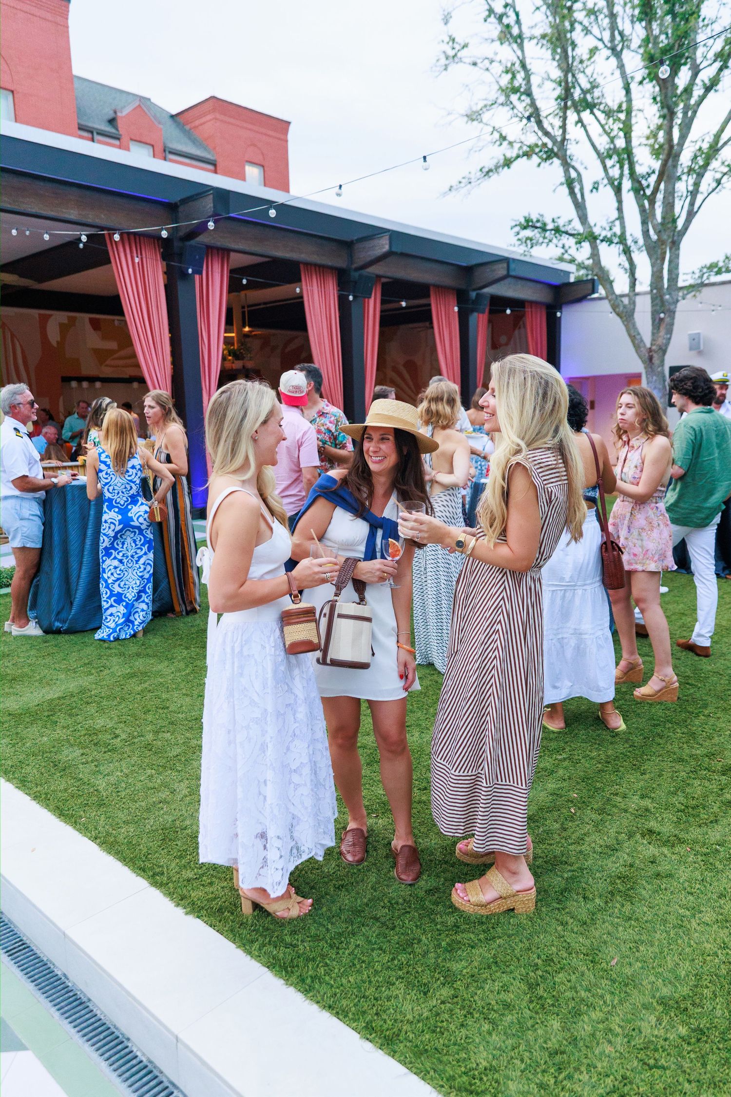 Three women in summer dresses socializing on a green lawn at an elegant outdoor party event.