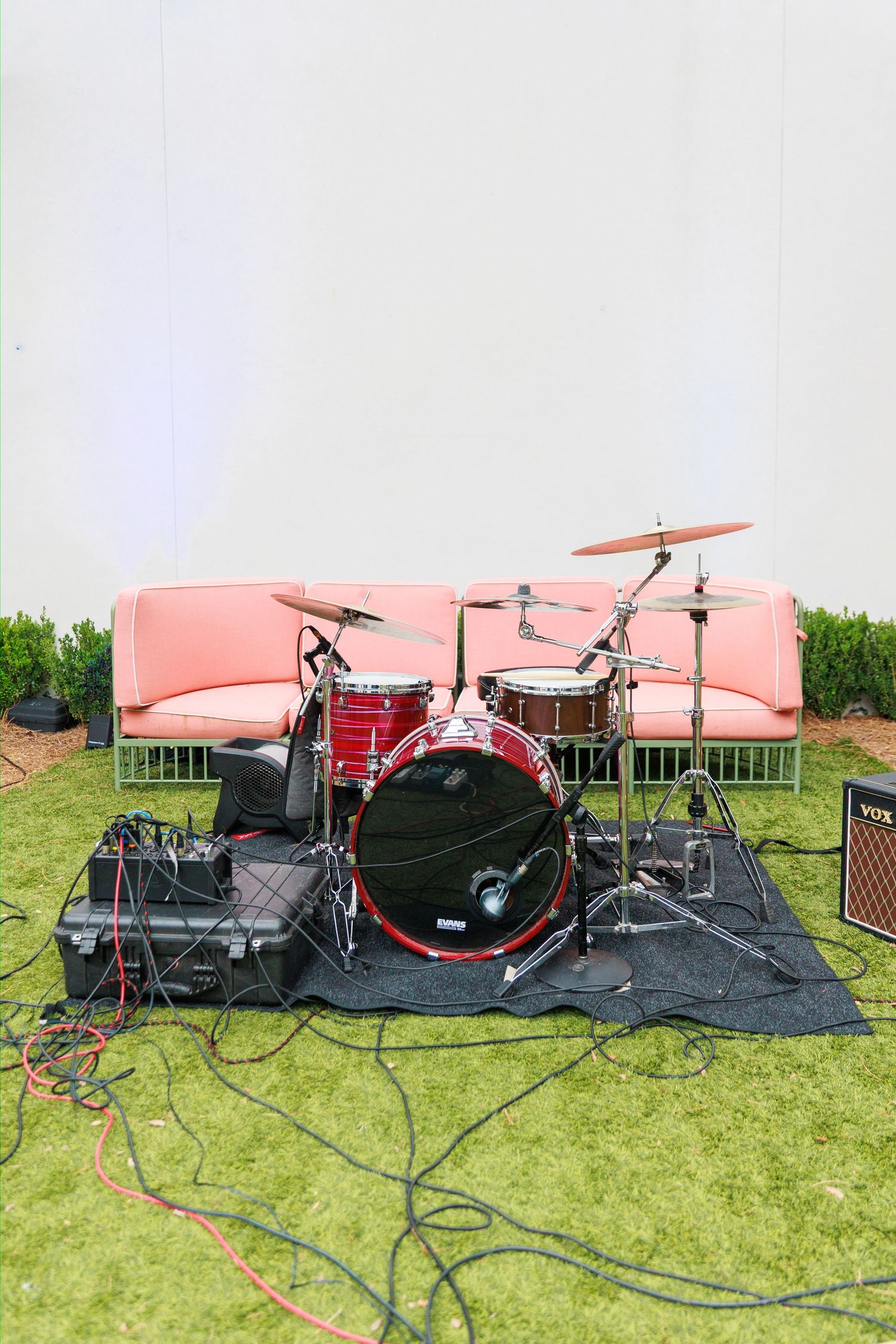 Red drum kit set up outdoors on grass in front of a pink sofa, surrounded by audio cables and equipment.